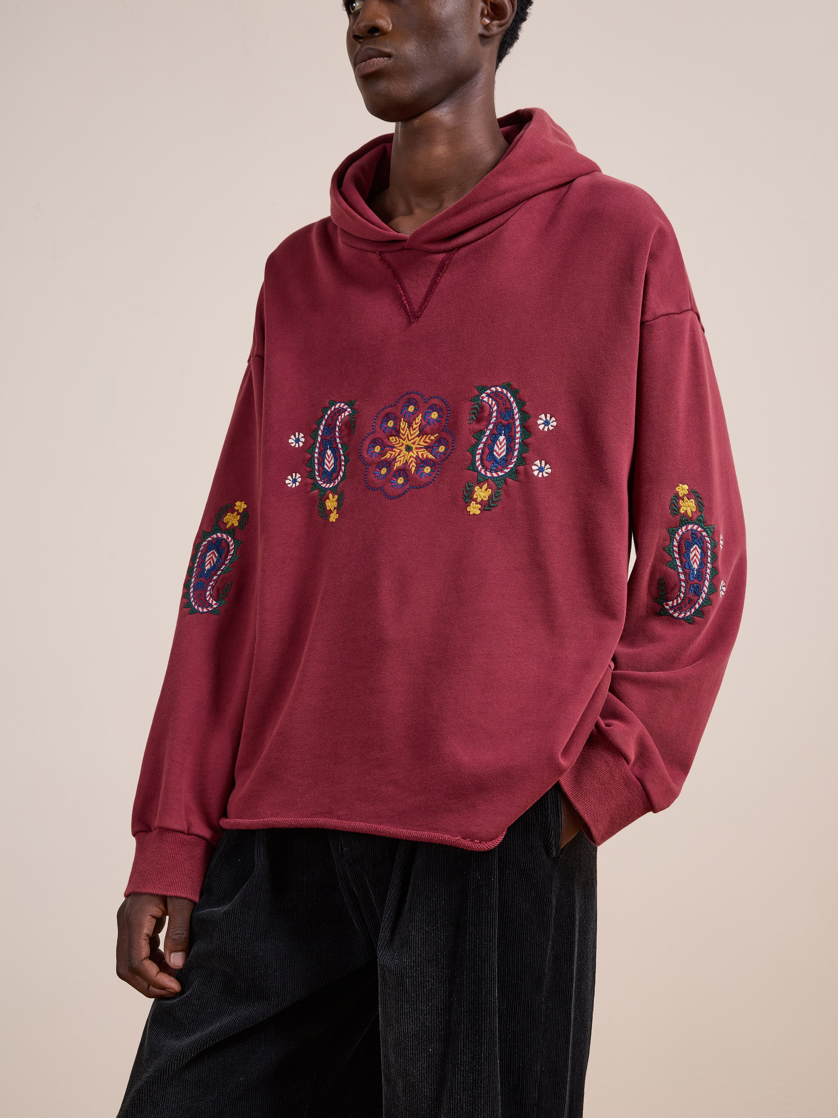A person wears the FOUND Embroidered Kantha Paisley Hoodie, a unisex relaxed-fit maroon cotton fleece with colorful Kantha-inspired paisley embroidery, paired with dark pants against a plain beige background.