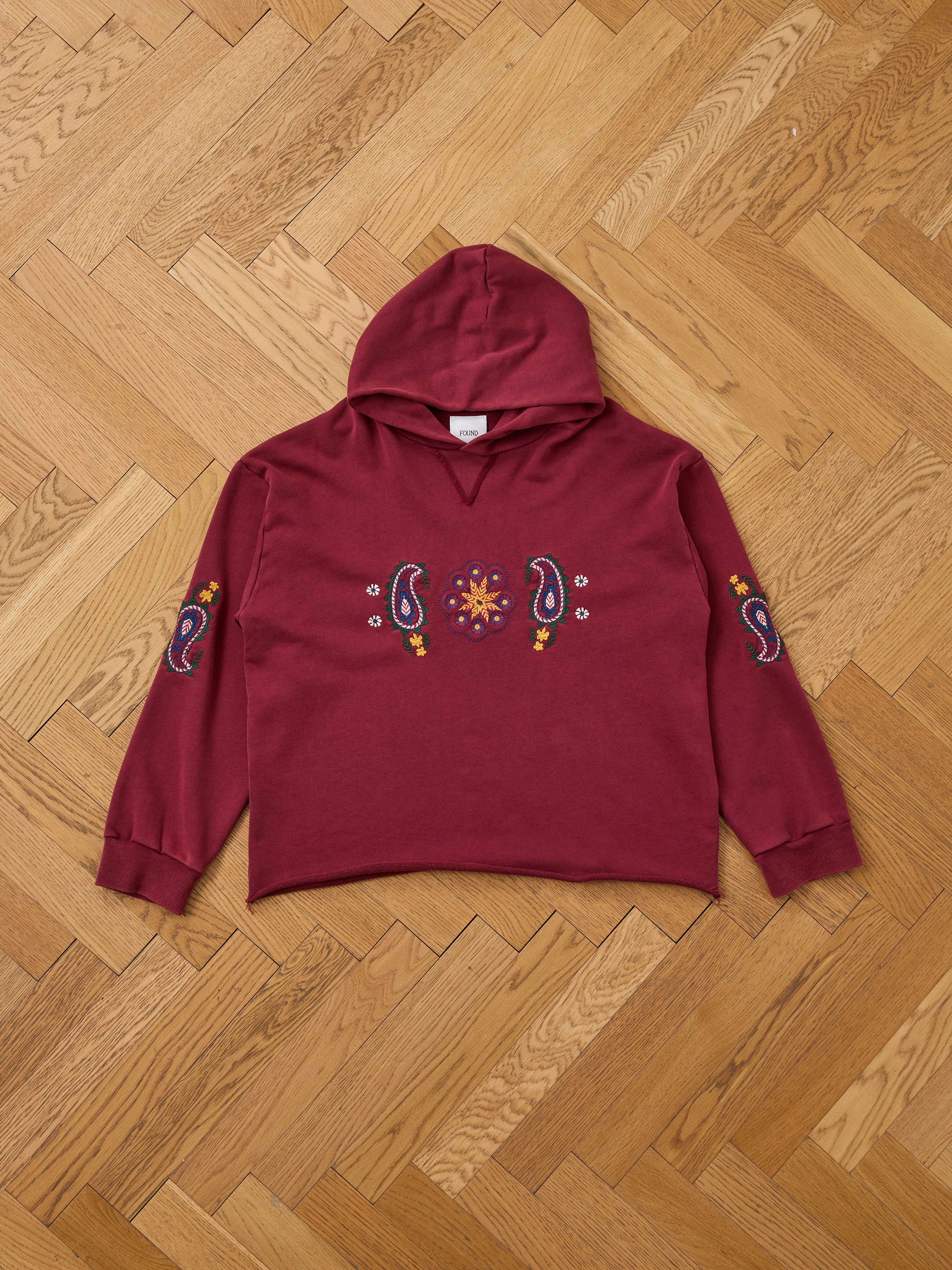 The FOUND Embroidered Kantha Paisley Hoodie in maroon cotton fleece features paisley embroidery and floral accents on the chest and sleeves, displayed flat on a light wooden herringbone floor.