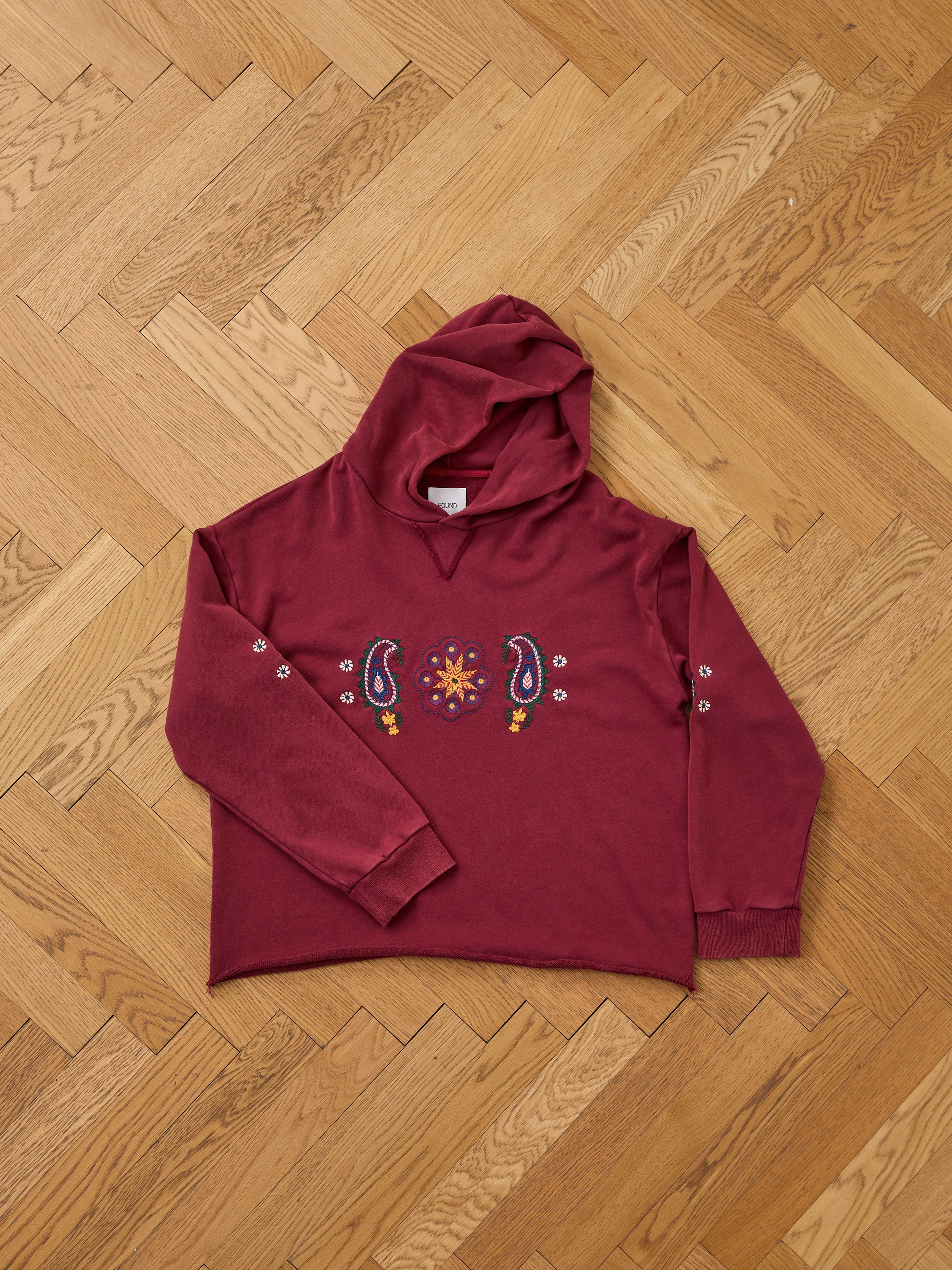 The FOUND Embroidered Kantha Paisley Hoodie, a maroon unisex cotton fleece with paisley embroidery on the chest and floral sleeve details, is shown laid flat on a light wooden herringbone floor.