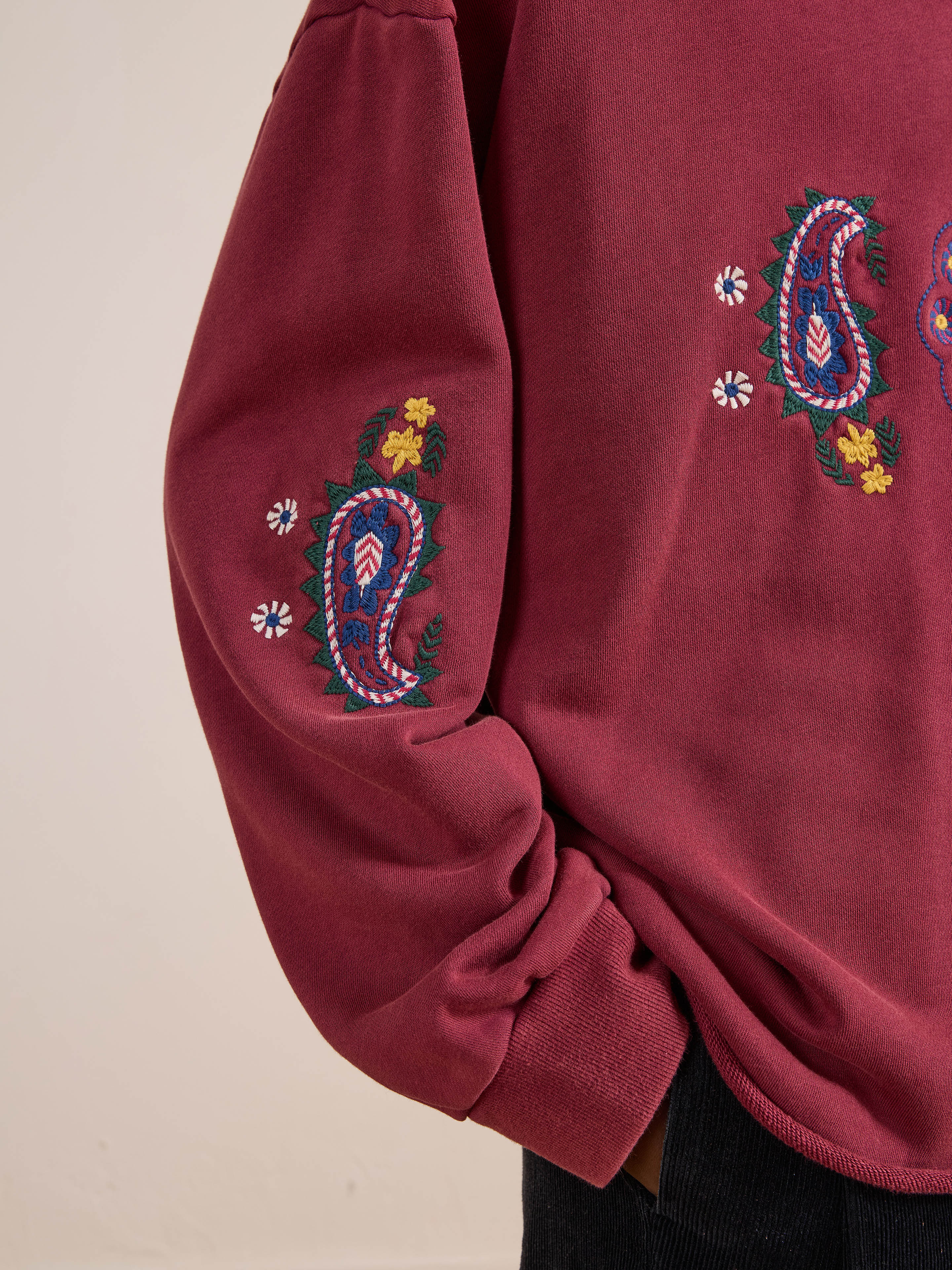 A person wears the FOUND Embroidered Kantha Paisley Hoodie, a maroon cotton fleece with paisley-inspired embroidery and colorful florals on the sleeve and chest.