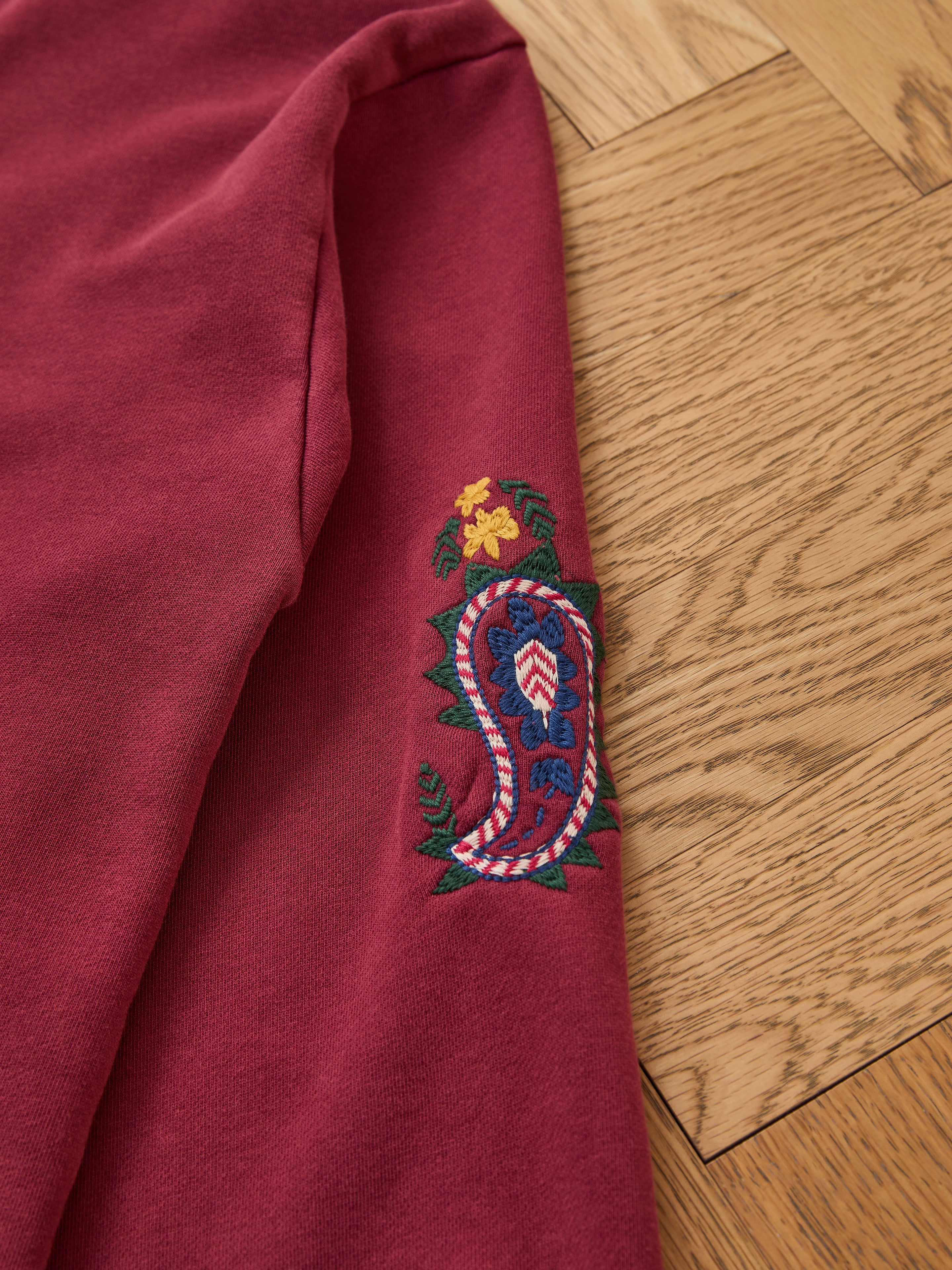 The FOUND Embroidered Kantha Paisley Hoodie is a maroon unisex relaxed fit cotton fleece with Kantha-inspired paisley embroidery and yellow flowers on the sleeve, displayed on a light wood floor.