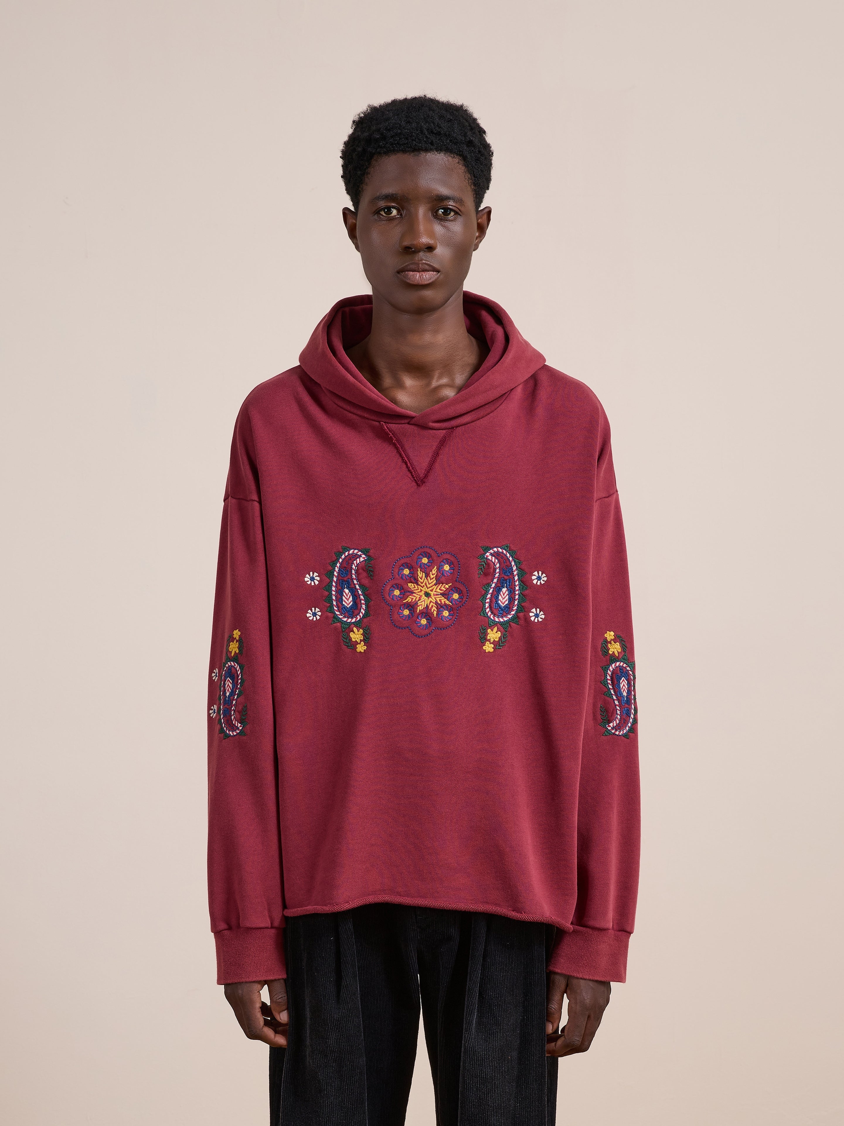 A person wearing the FOUND Embroidered Kantha Paisley Hoodie—a loose, unisex relaxed fit hoodie adorned with colorful Kantha embroidery on the front and sleeves—stands against a plain beige background.