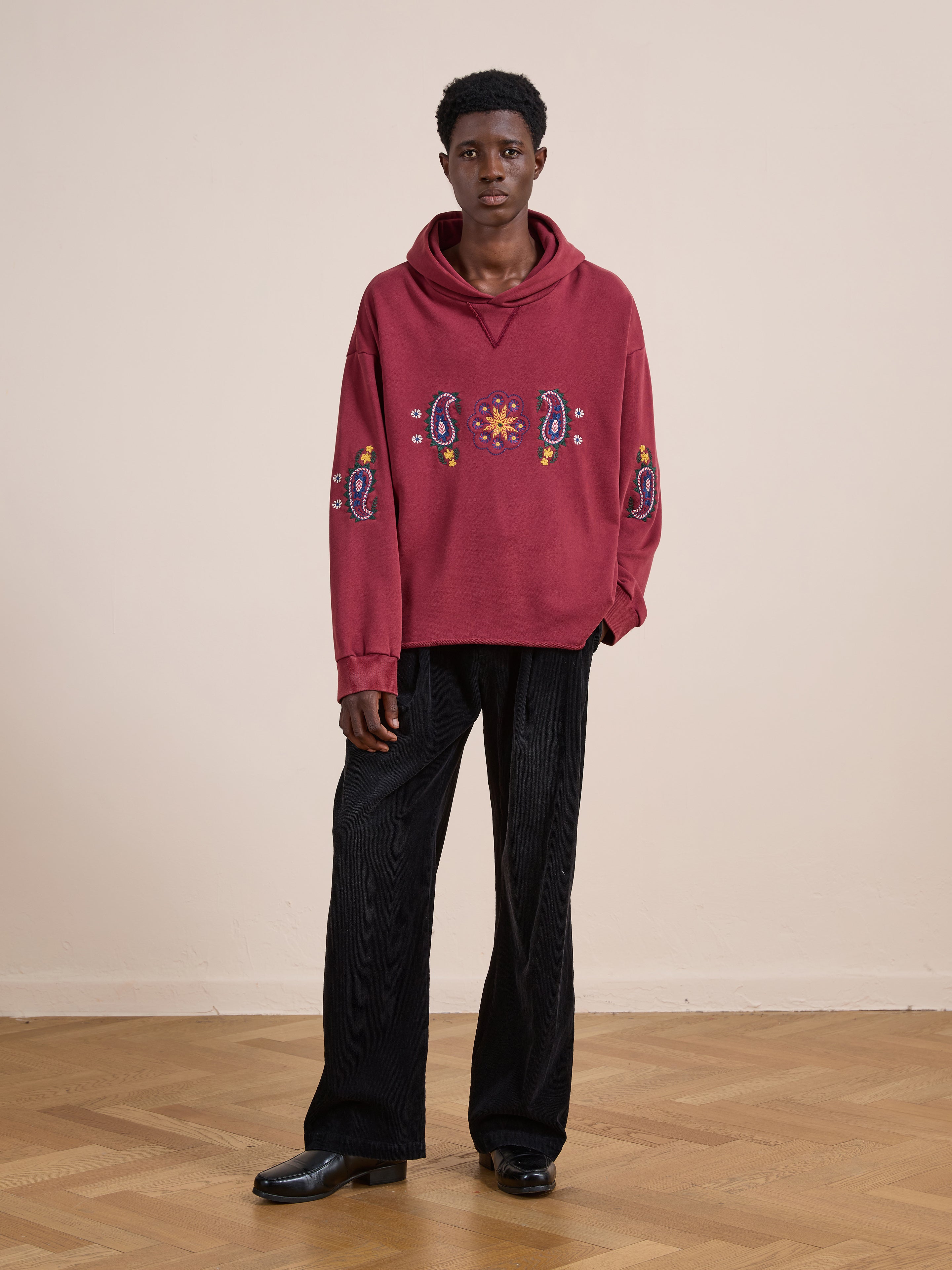 Indoors, a person wears the FOUND Embroidered Kantha Paisley Hoodie in maroon cotton fleece with paisley embroidery, paired with unisex relaxed fit black wide-leg pants and black shoes, set against a light wall and wooden floor.