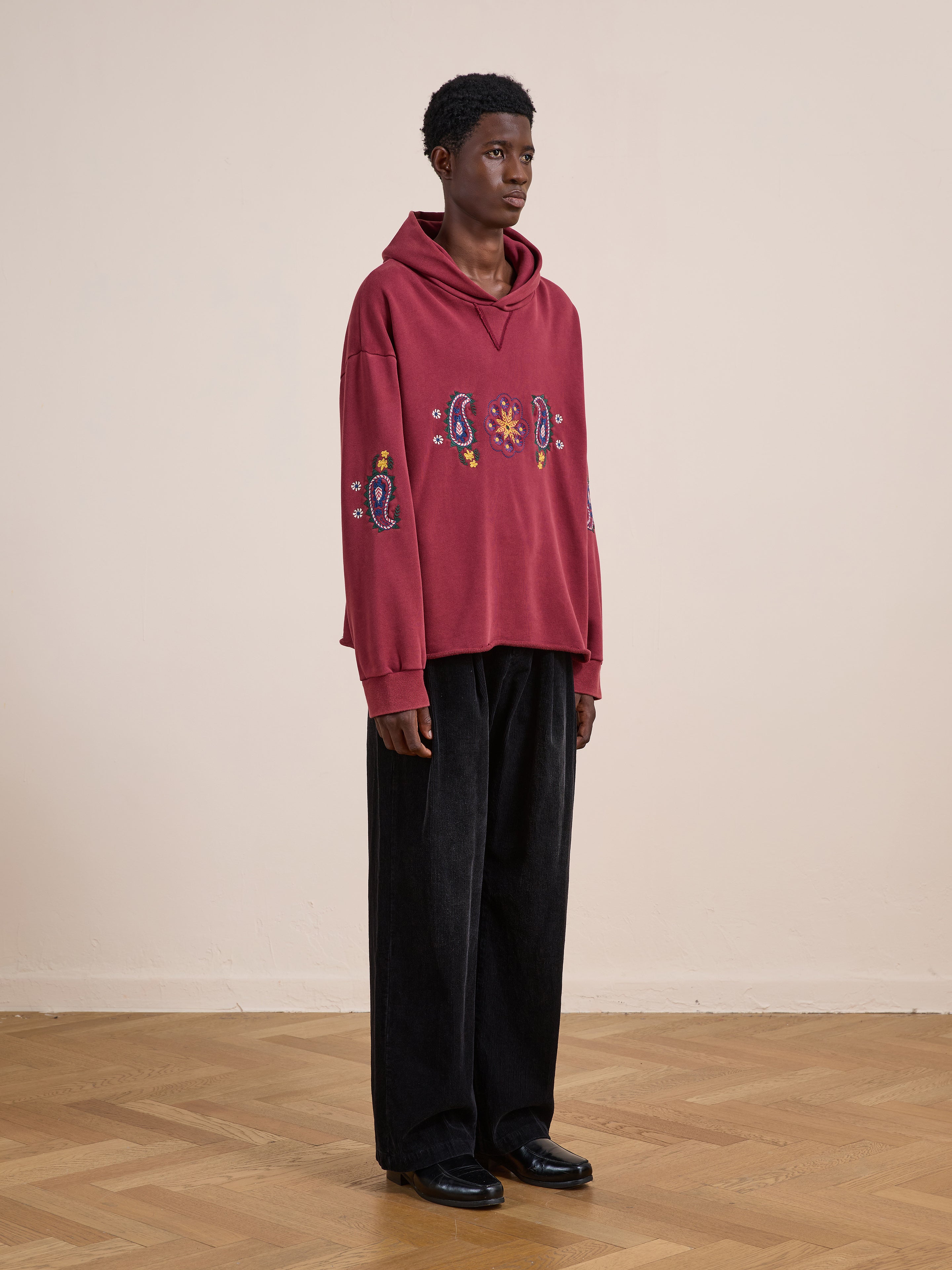 A person wears the FOUND Embroidered Kantha Paisley Hoodie—a unisex relaxed fit cotton fleece with paisley and sun embroidery—paired with black wide-leg pants and shoes, standing on a wooden floor against a beige wall.