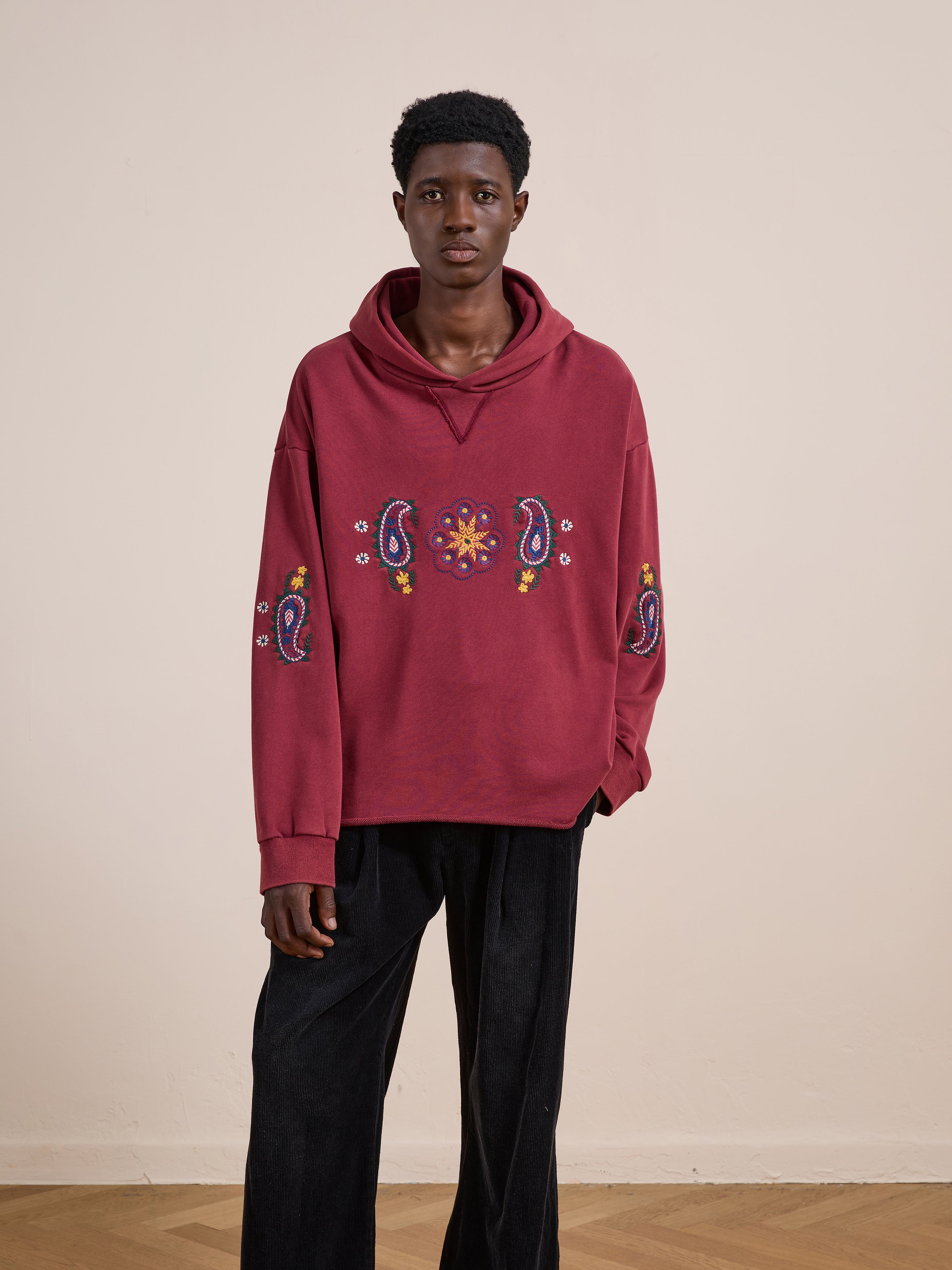 A person indoors wears the FOUND Embroidered Kantha Paisley Hoodie in red cotton fleece with paisley embroidery, paired with black pants, against a plain beige wall.