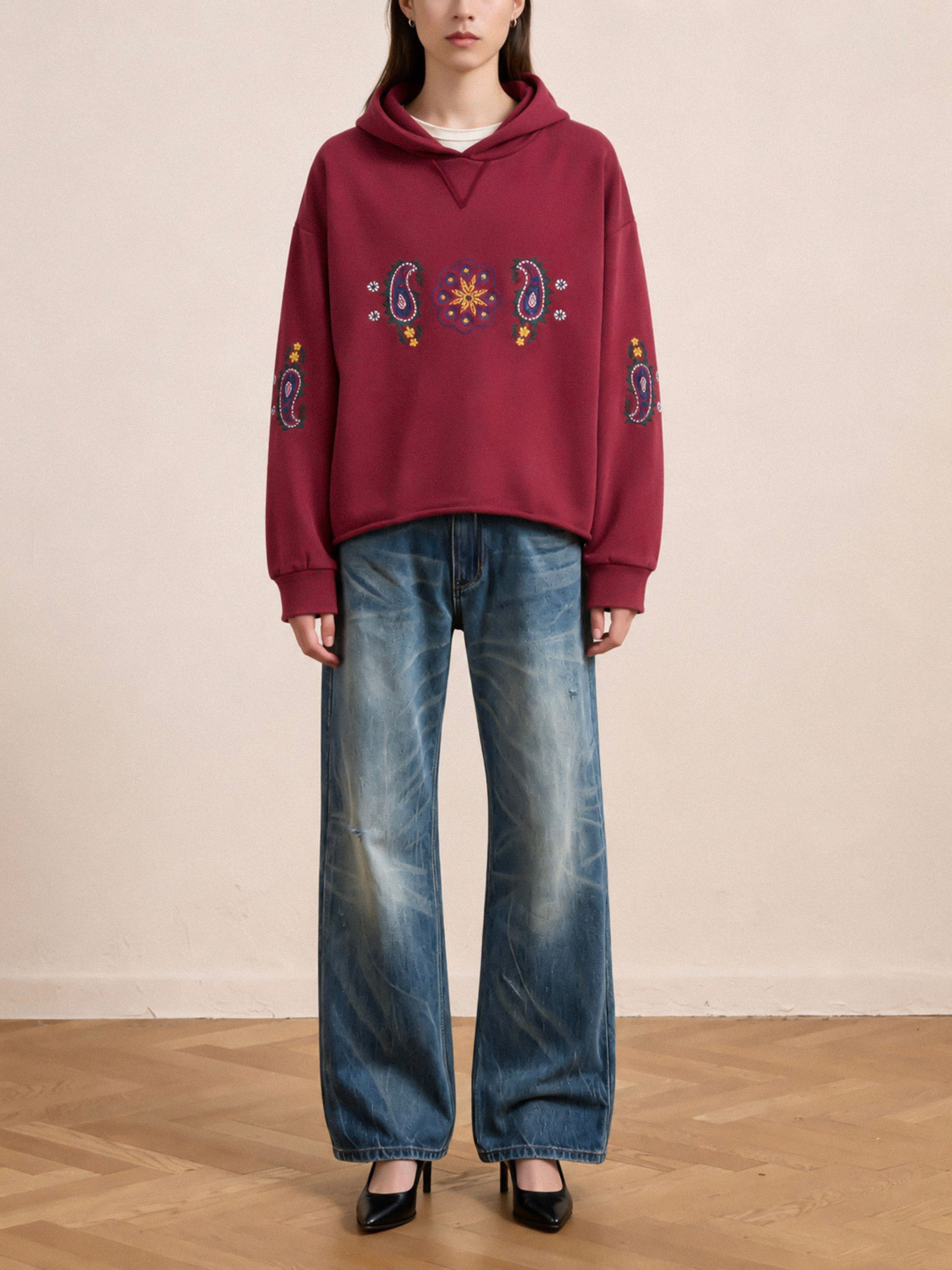 A person is standing indoors on a wooden floor, wearing the FOUND Embroidered Kantha Paisley Hoodie featuring intricate maroon paisley embroidery, paired with loose faded blue jeans and black high-heeled shoes.