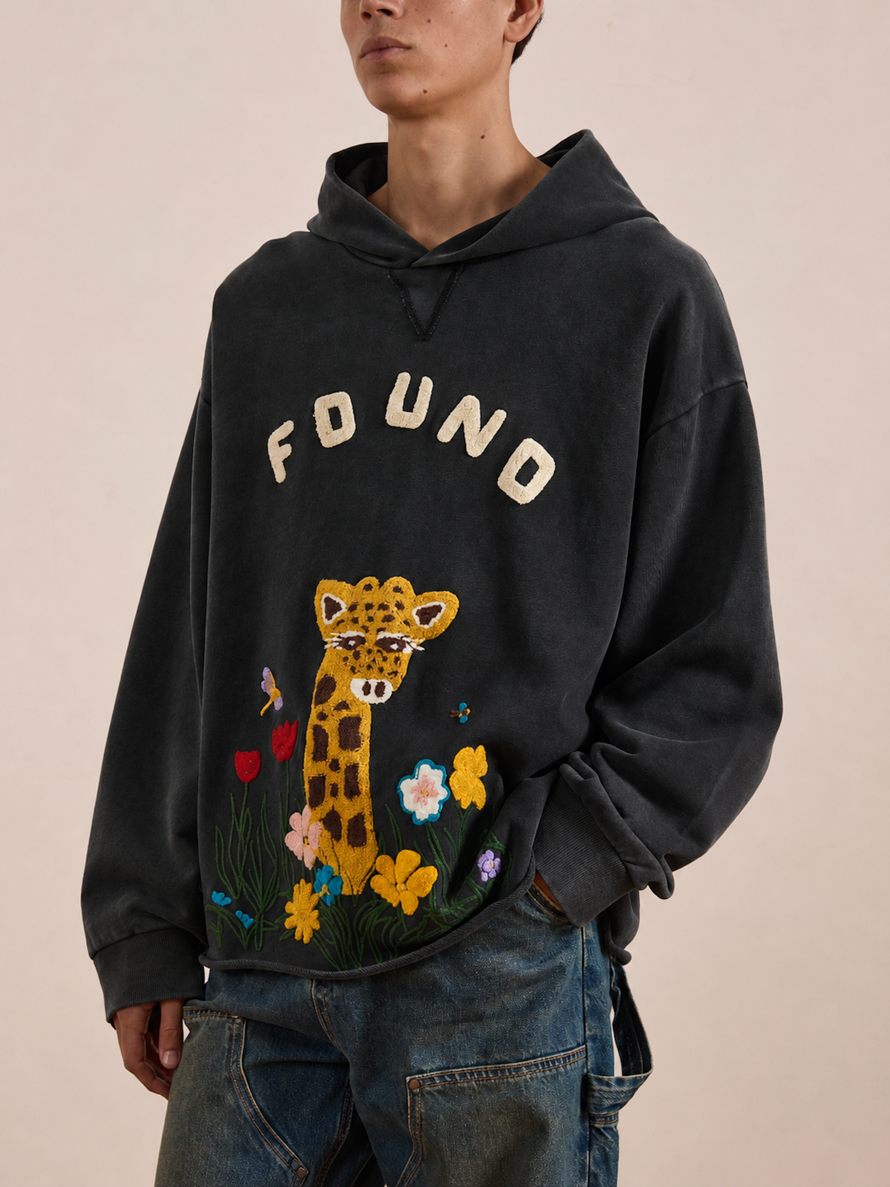 A person wearing the FOUND Embroidered Wildlife Hoodie—featuring “FOUND” text and an embroidered giraffe surrounded by flowers and butterflies on the front, crafted from vintage French-terry cotton—styled with blue jeans.