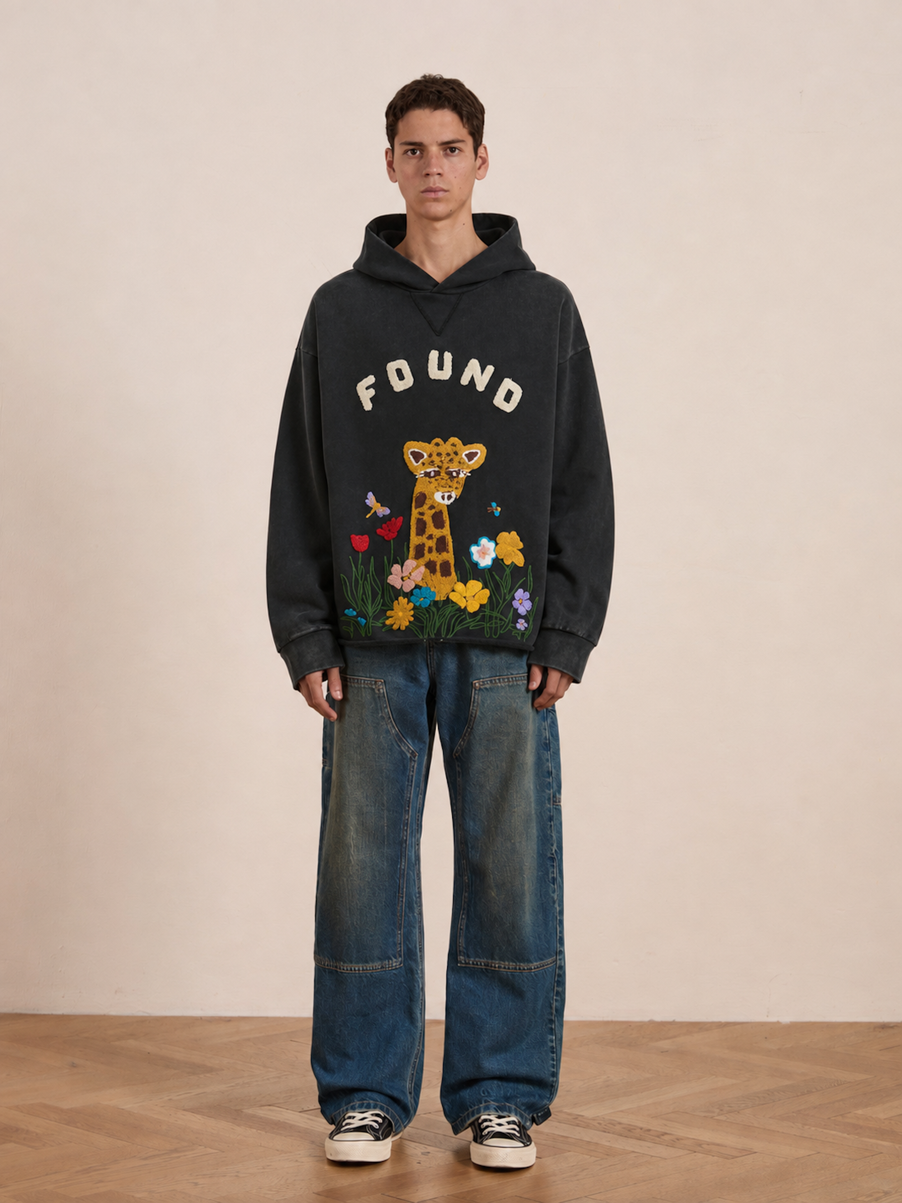 A person wears the FOUND Embroidered Wildlife Hoodie in black, featuring “FOUND” text and a cartoon giraffe amid flowers, crafted from vintage French-terry cotton. They pair it with baggy blue jeans and black sneakers, standing on a wooden floor against a plain wall.