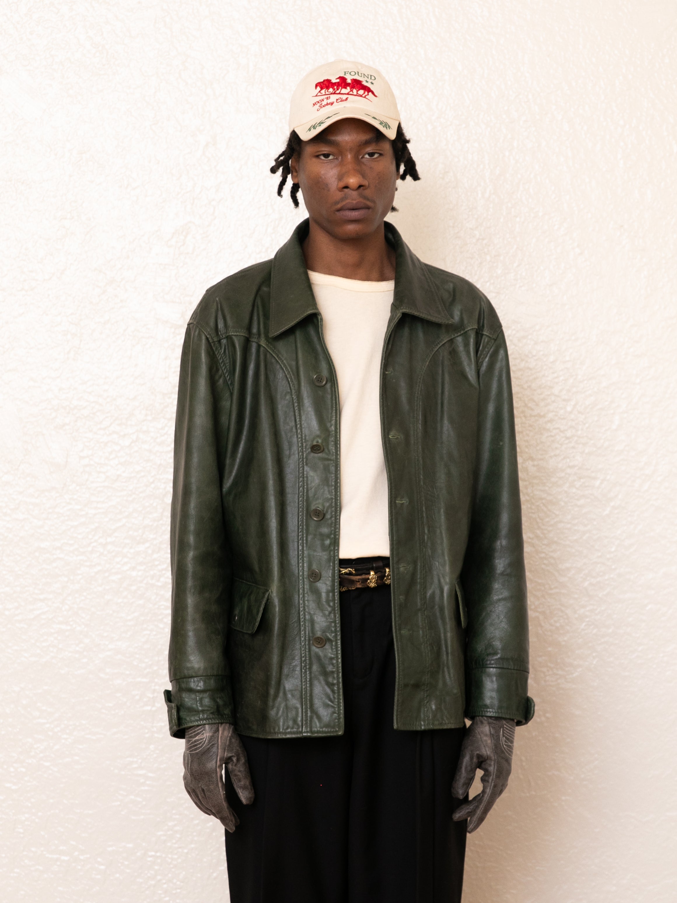 A person wearing a cream baseball cap, off-white shirt, FOUND Leather Field Jacket in emerald green, black pants, and gray gloves stands against a plain white textured wall.
