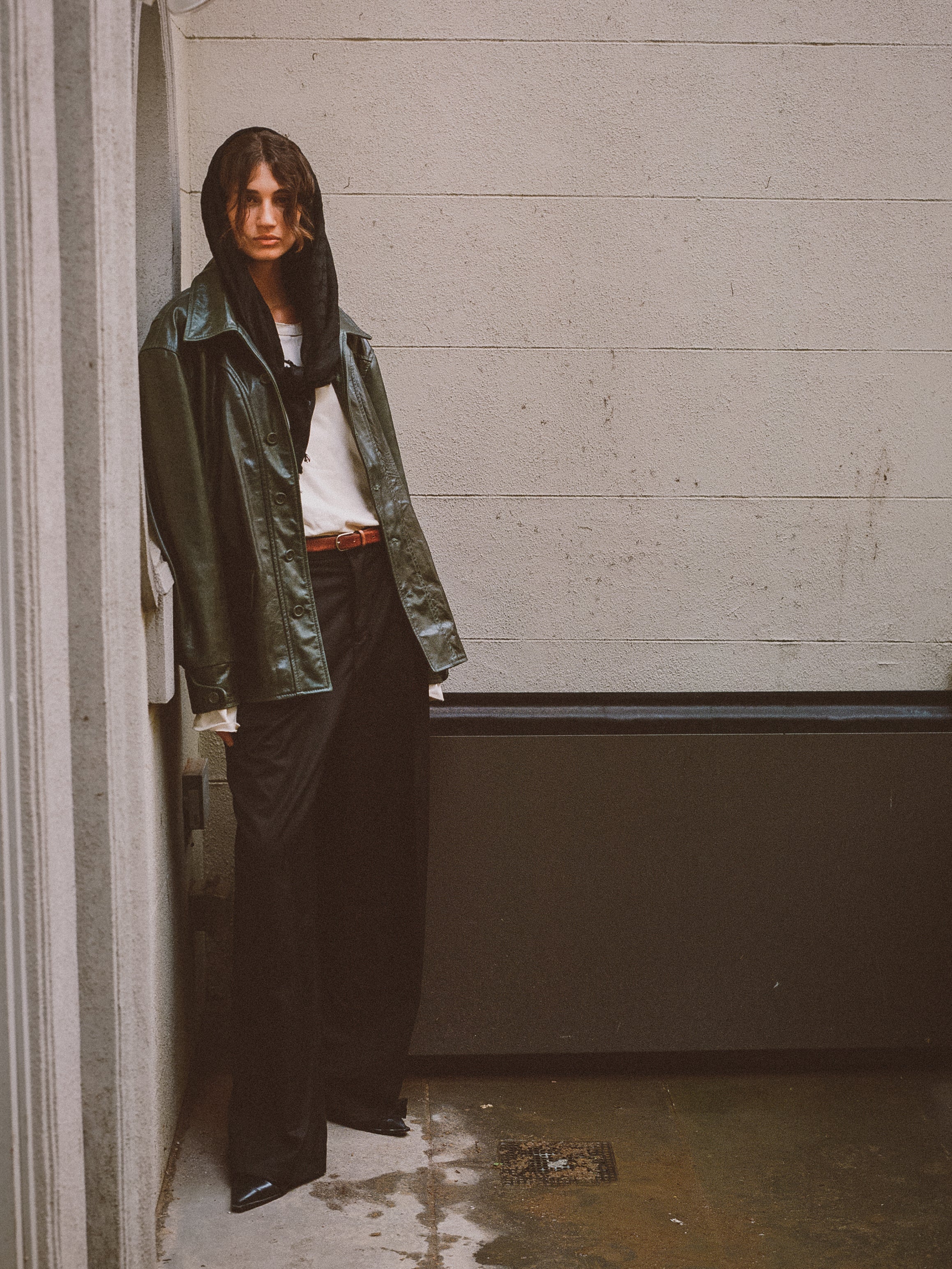 A person in a FOUND Leather Field Jacket—emerald green, hand-distressed leather—paired with a black hood, white shirt, black pants, and black shoes stands against a light wall in a narrow outdoor space.