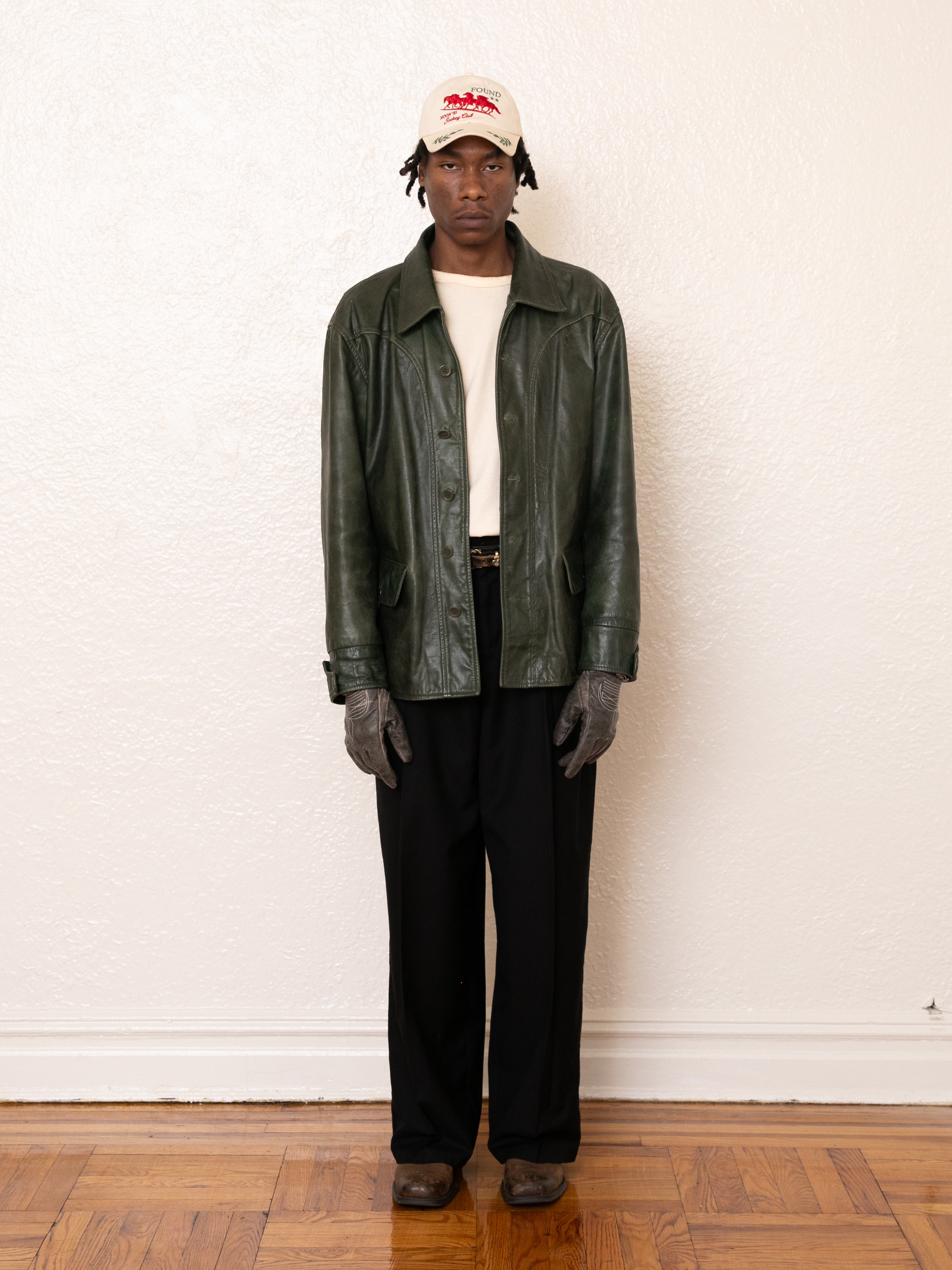 A person stands indoors against a plain wall, dressed in FOUND's emerald green Leather Field Jacket, paired with a white shirt, dark pants, gray gloves, brown shoes, and a beige cap.
