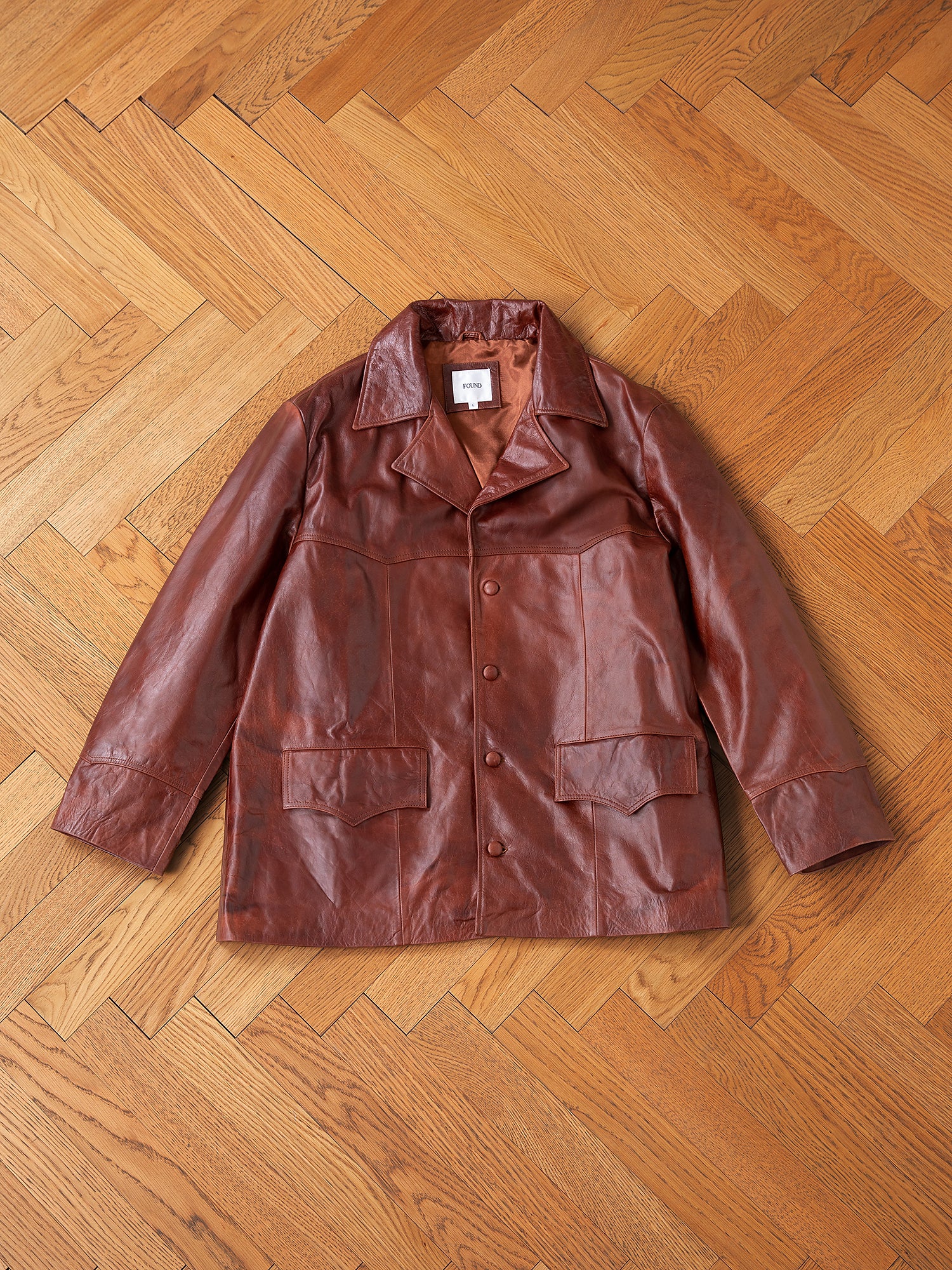 The Claret Western Oversized Leather Blazer from FOUND, featuring front buttons and large pockets, is strikingly displayed on a wooden herringbone-patterned floor.