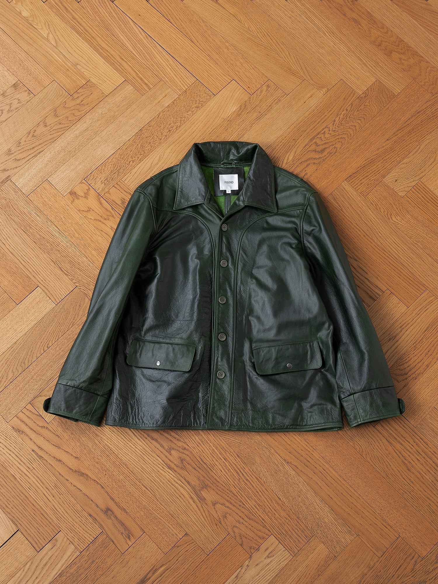 The FOUND Leather Field Jacket in emerald green hand-distressed leather, featuring buttons and flap pockets, is laid flat on a light brown wooden herringbone floor.