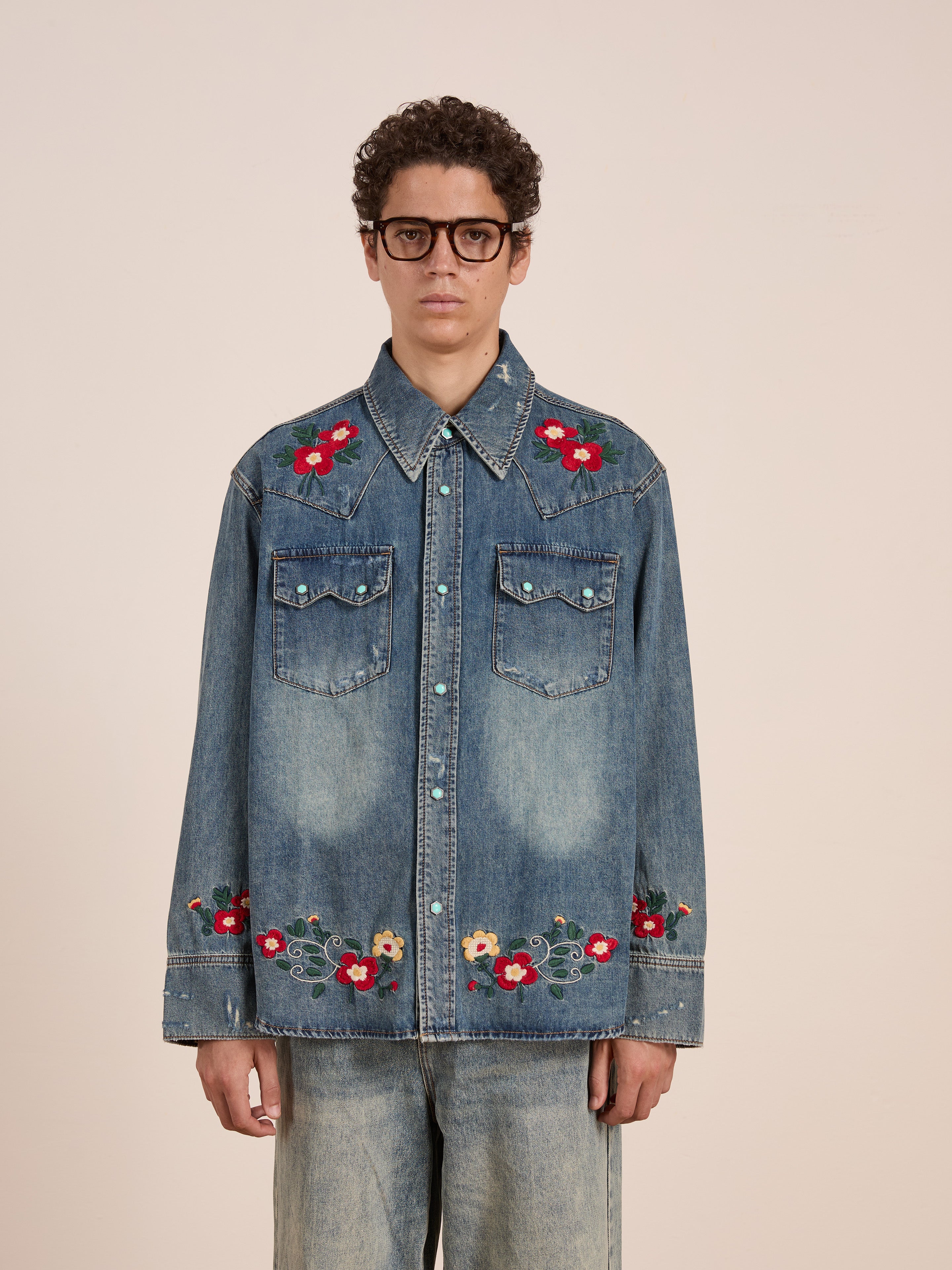 A person in glasses wears the FOUND Floral Embroidered Western Denim Shirt with floral details on the collar, chest, and cuffs, paired with light-wash jeans, standing against a plain background.