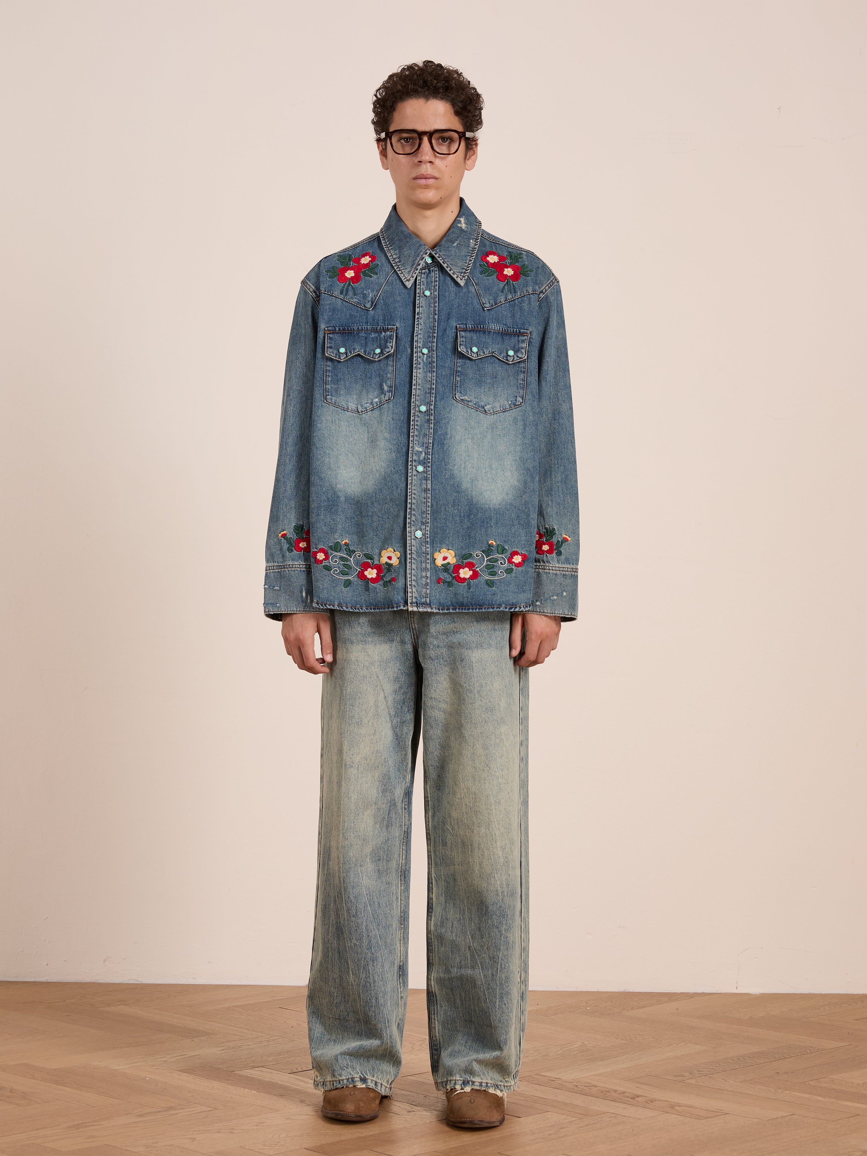 Indoors, a person wears the FOUND Floral Embroidered Western Denim Shirt with red floral details, matching wide-leg jeans in structured denim, brown shoes, and oversized glasses.