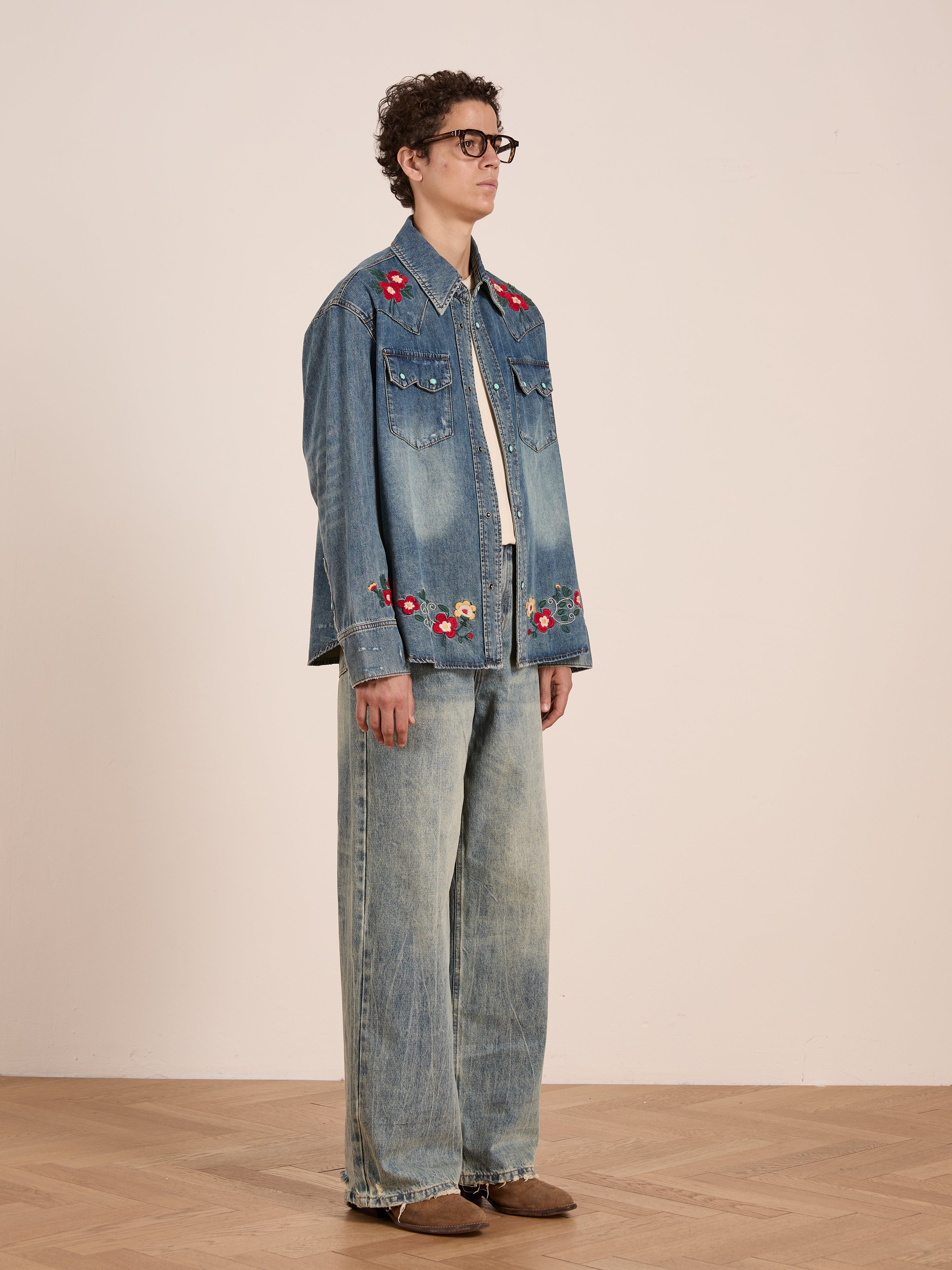 A person wearing glasses, the FOUND Floral Embroidered Western Denim Shirt, wide-leg jeans, and brown shoes stands against a plain light-colored wall on a wooden floor.