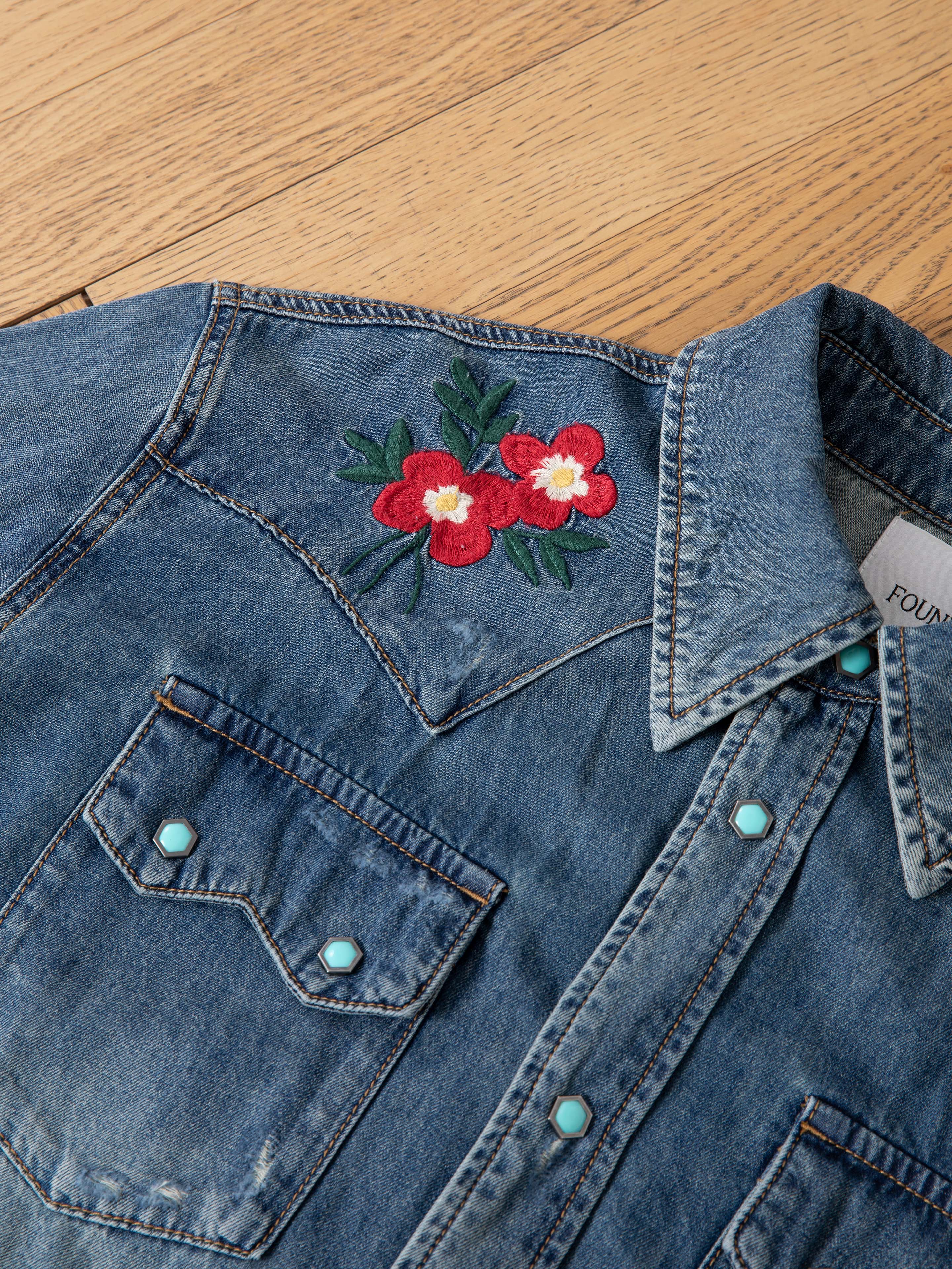 The FOUND Floral Embroidered Western Denim Shirt, with turquoise snaps and vivid red floral embroidery on the shoulders, is laid flat on a wooden surface.