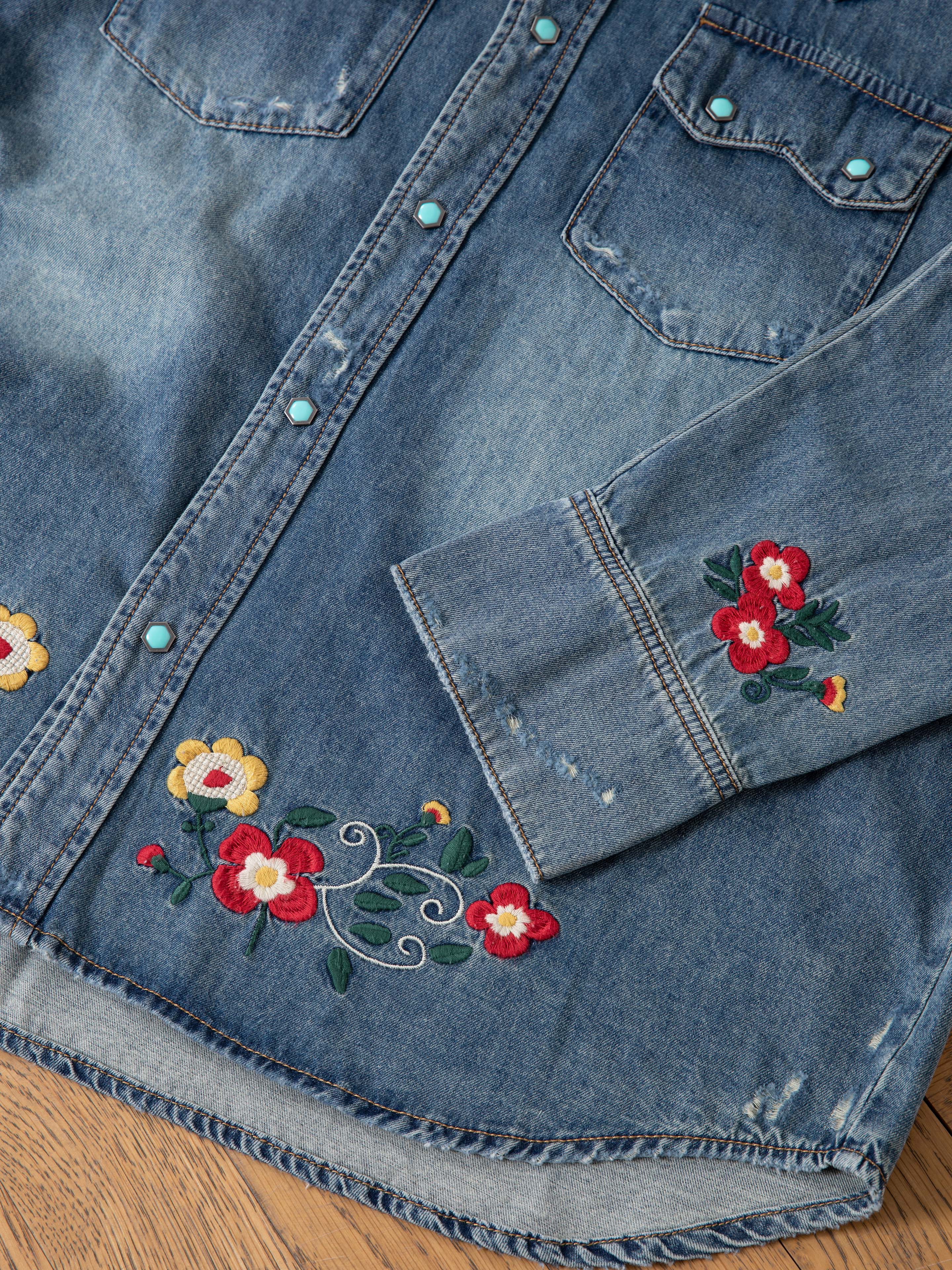 The FOUND Floral Embroidered Western Denim Shirt features vibrant red and yellow floral embroidery on the sleeve, pocket, and hem, shown laid flat on a wooden surface.