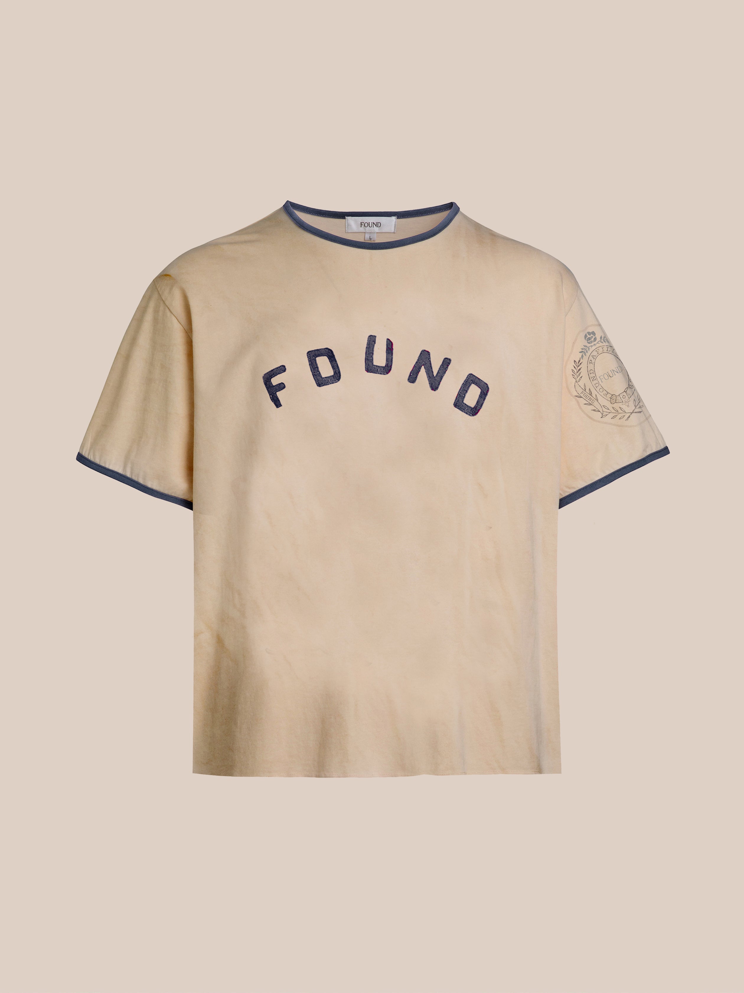 The FOUND Logo Felt Patch Ringer Tee is a beige unisex relaxed fit tee with a navy collar and sleeve trim, "FOUND" in navy on the chest, a graphic on the left sleeve—perfect for everyday wear.