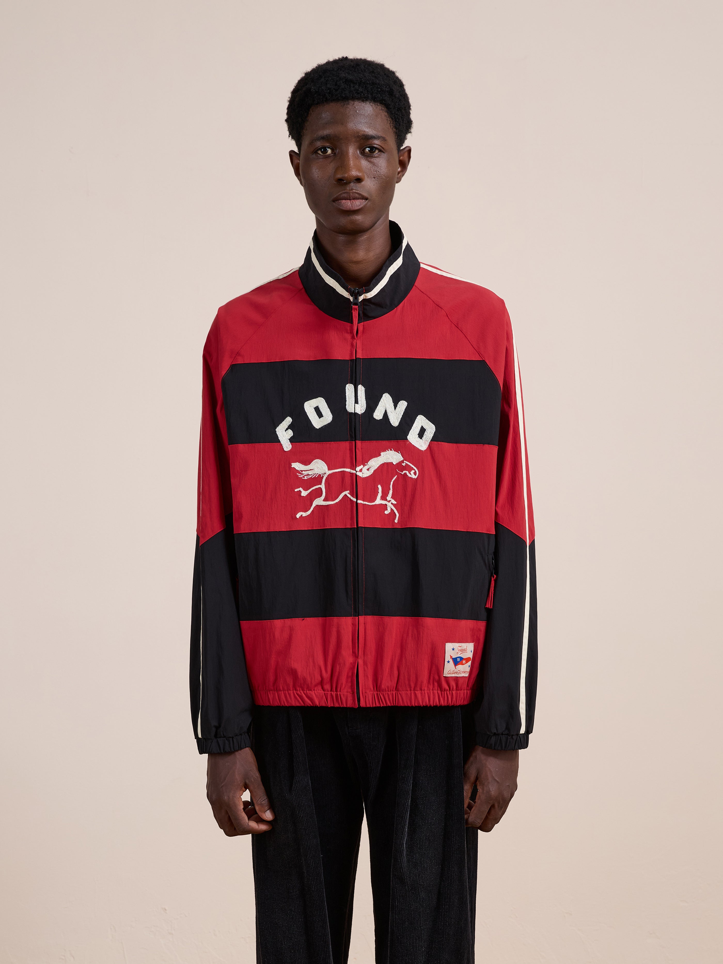 A person in a FOUND Racing Striped Windbreaker featuring "FOUND" and a running animal graphic on the front, styled like vintage sportswear, stands against a plain background.