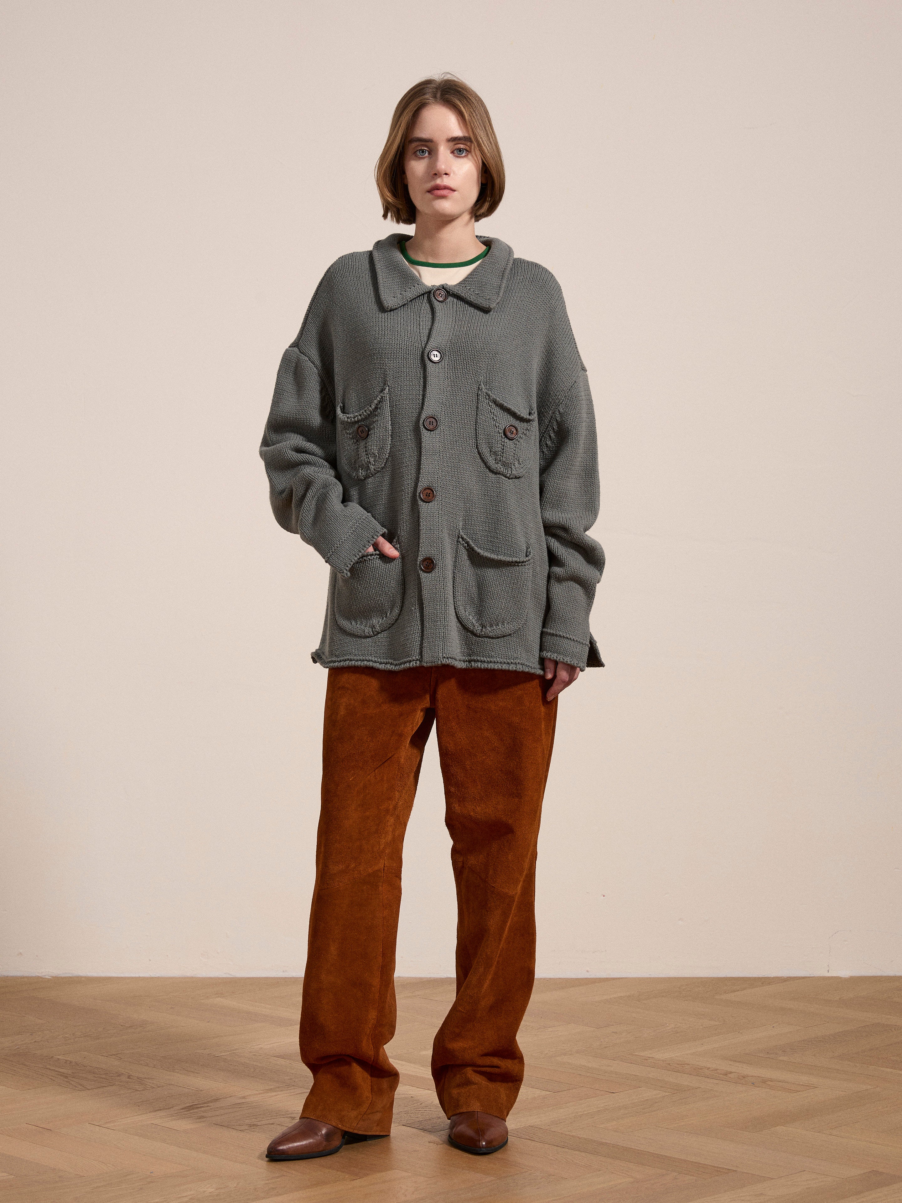 A person wearing a cozy, oversized gray Hunter Knit Collared Cardigan by FOUND, paired with brown pants and shoes, stands on a wooden floor against a plain beige wall.
