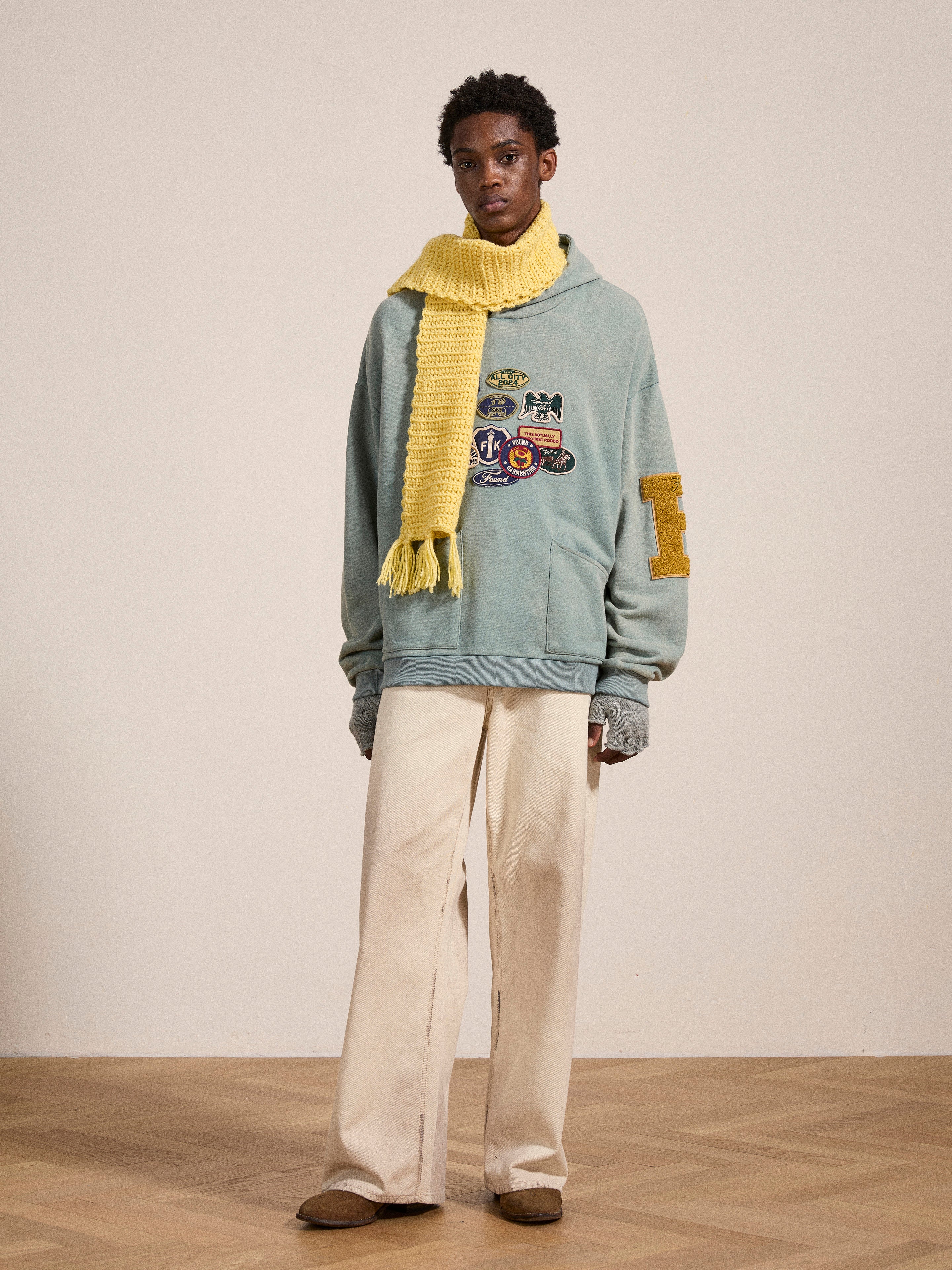 A person stands on a wooden floor wearing a Multi Patch Hoodie by FOUND made of French Terry cotton, paired with a yellow knit scarf, beige pants, and brown shoes. The background is a plain, light-colored wall.