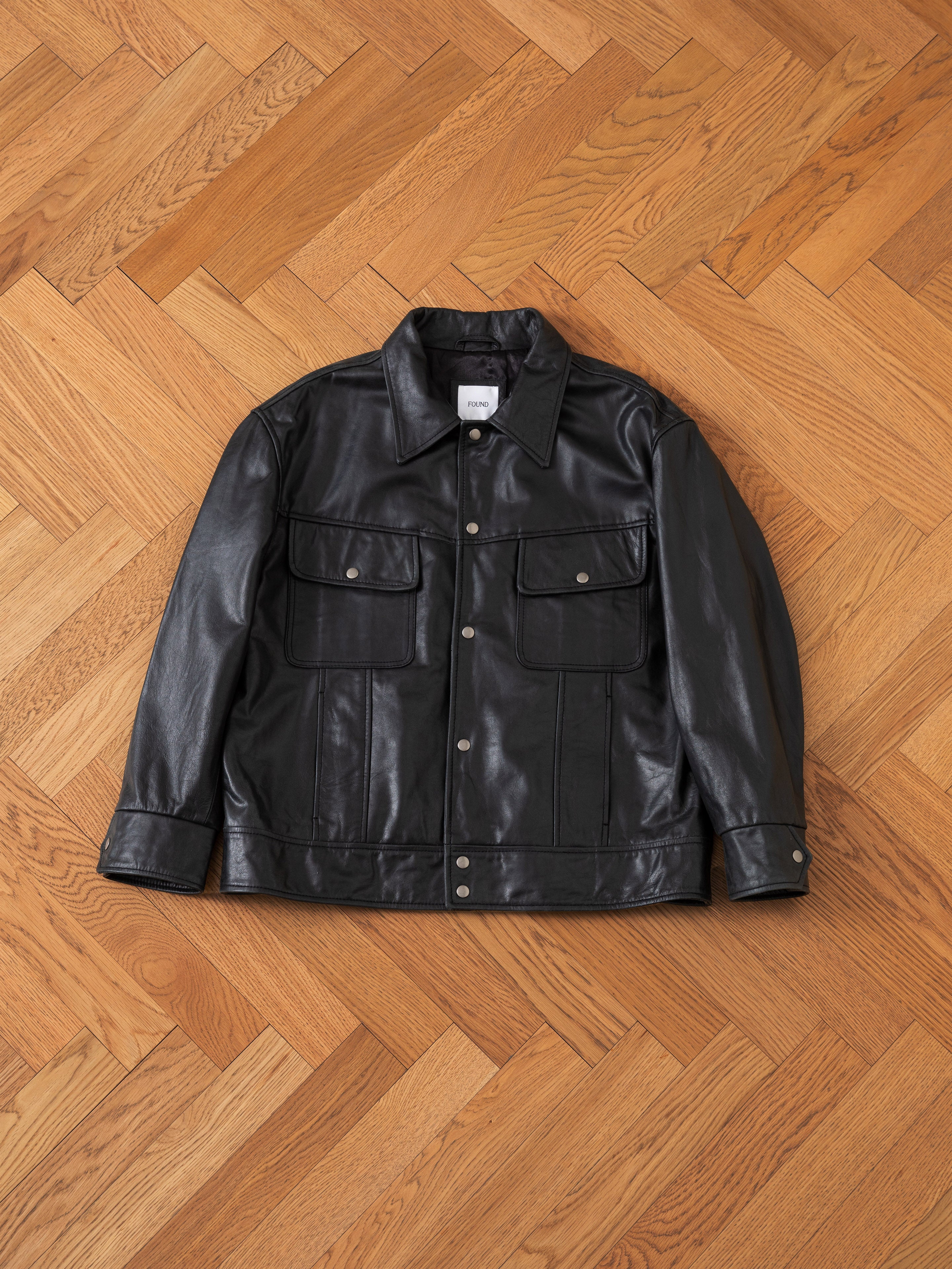 A FOUND Leather Trucker Jacket in black, featuring a button-up front and two chest pockets, is laid flat on a wooden herringbone-patterned floor.