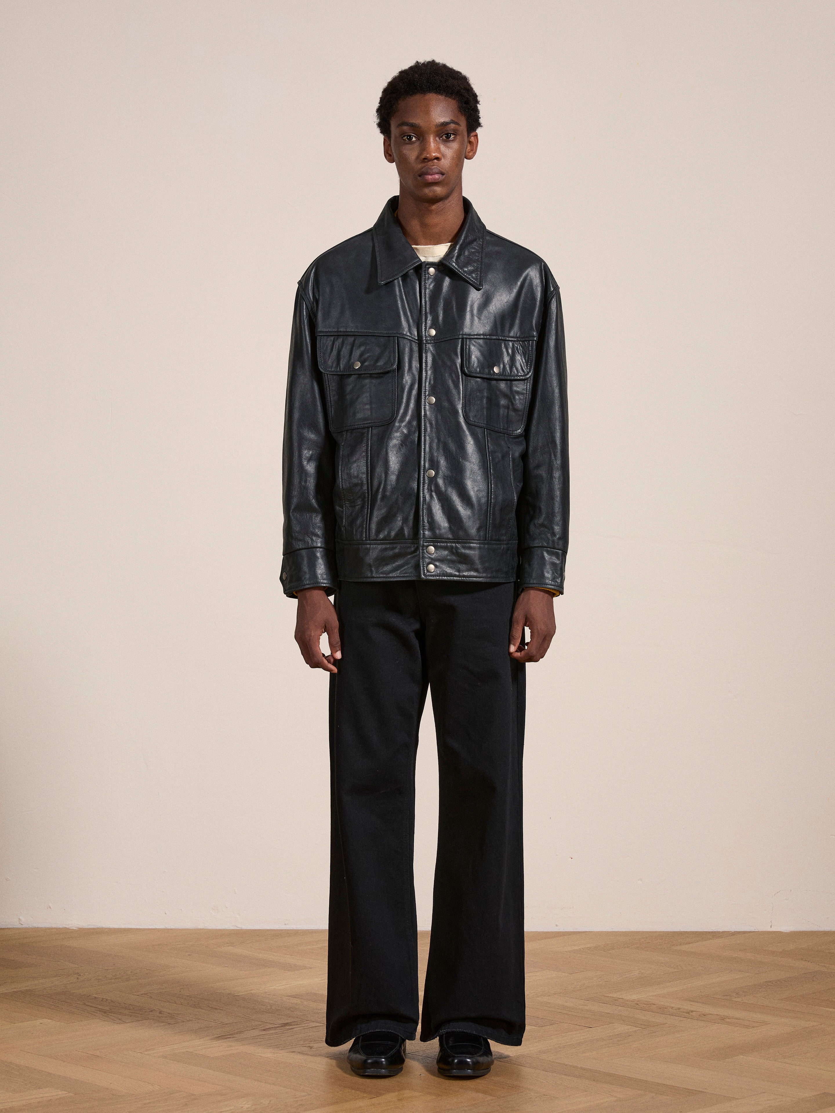 A person stands in a room wearing the FOUND Leather Trucker Jacket, which is classic black with multiple pockets, paired with black wide-leg pants. The room features a plain, light-colored wall and a wooden floor.
