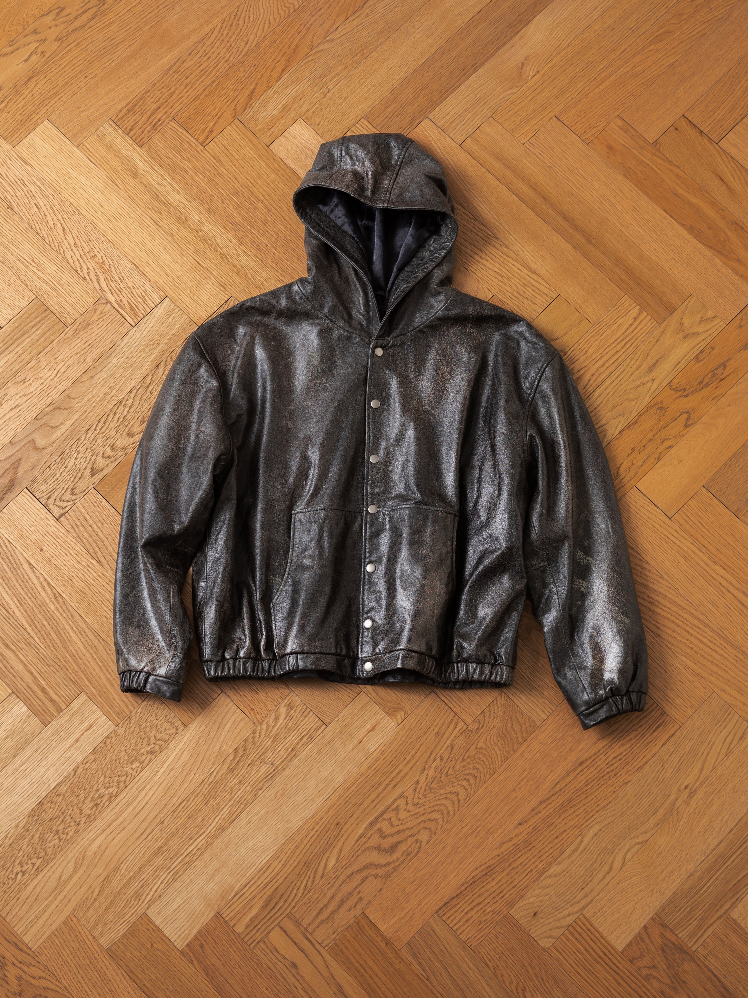 The Distressed Genuine Leather Hoodie by Found, crafted from lightweight cowhide in a metallic silver finish and featuring snap buttons along with elastic cuffs and hem, is laid flat on a wooden parquet floor.