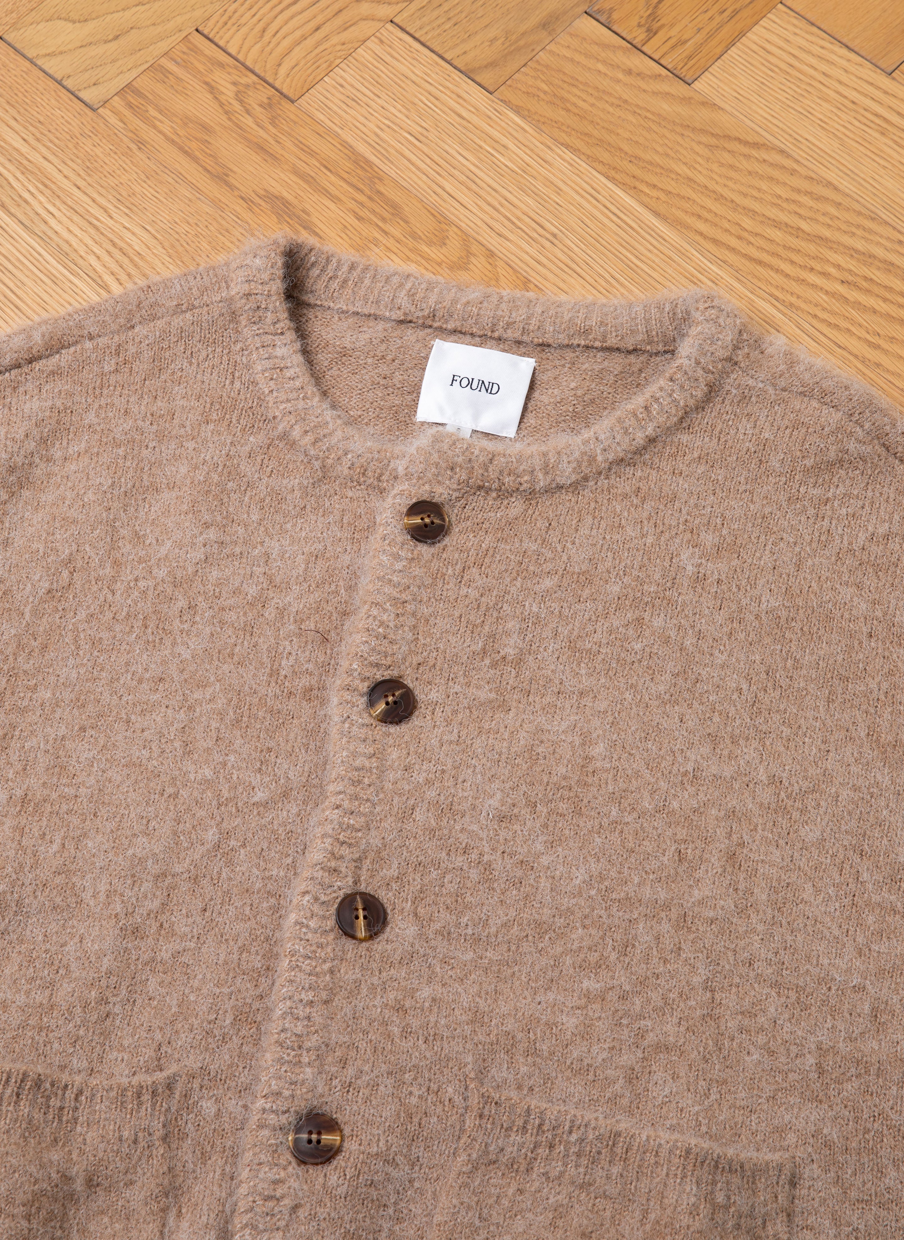 A beige mohair cardigan with tortoise buttons is laid flat on a wooden herringbone floor. A label inside the collar reads "FOUND.