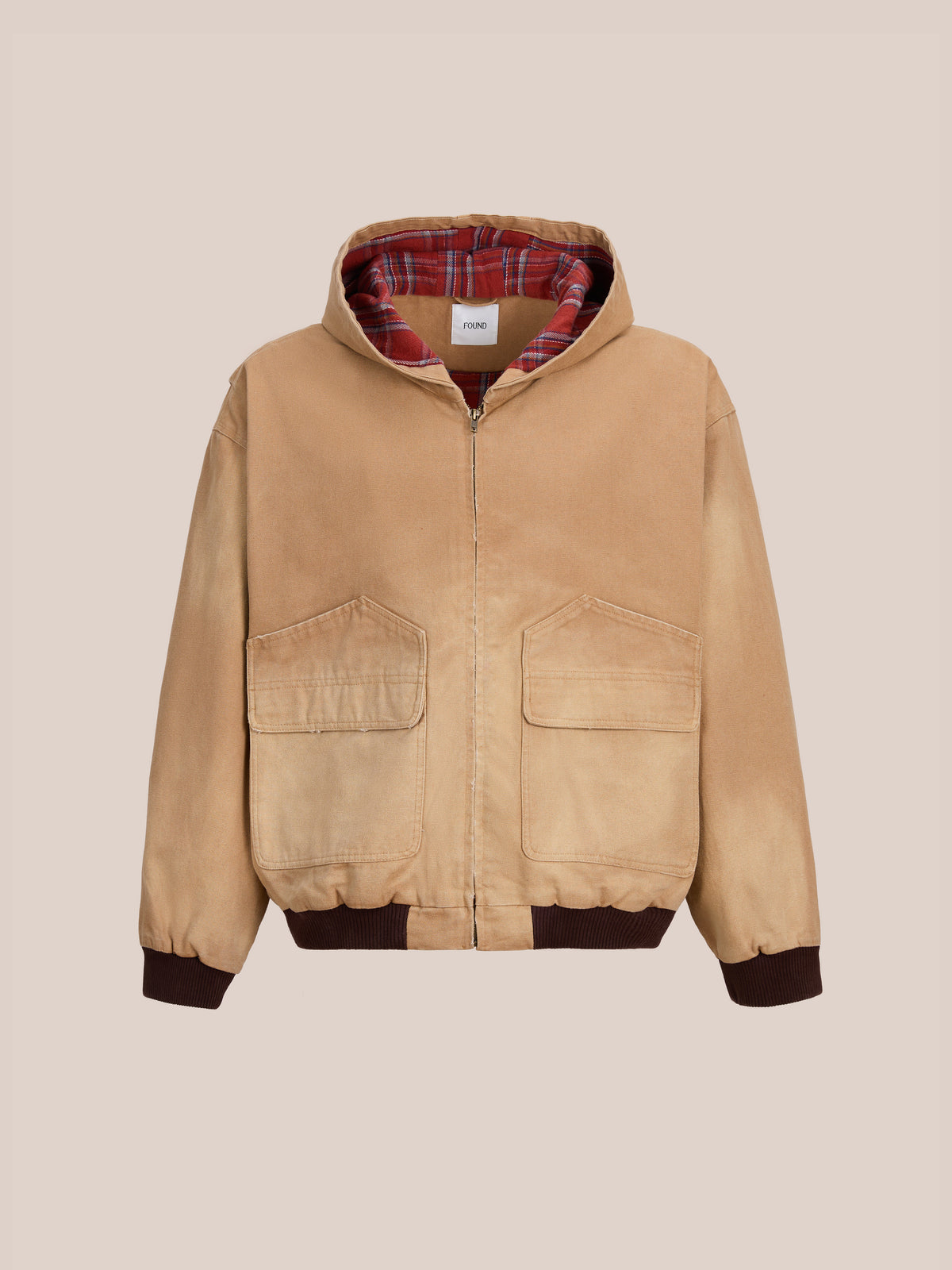 The FOUND Faded Canvas Work Hoodie is a tan unisex zip-up with two large flap pockets, ribbed cuffs and hem, and red plaid lining, shown on a light beige background.