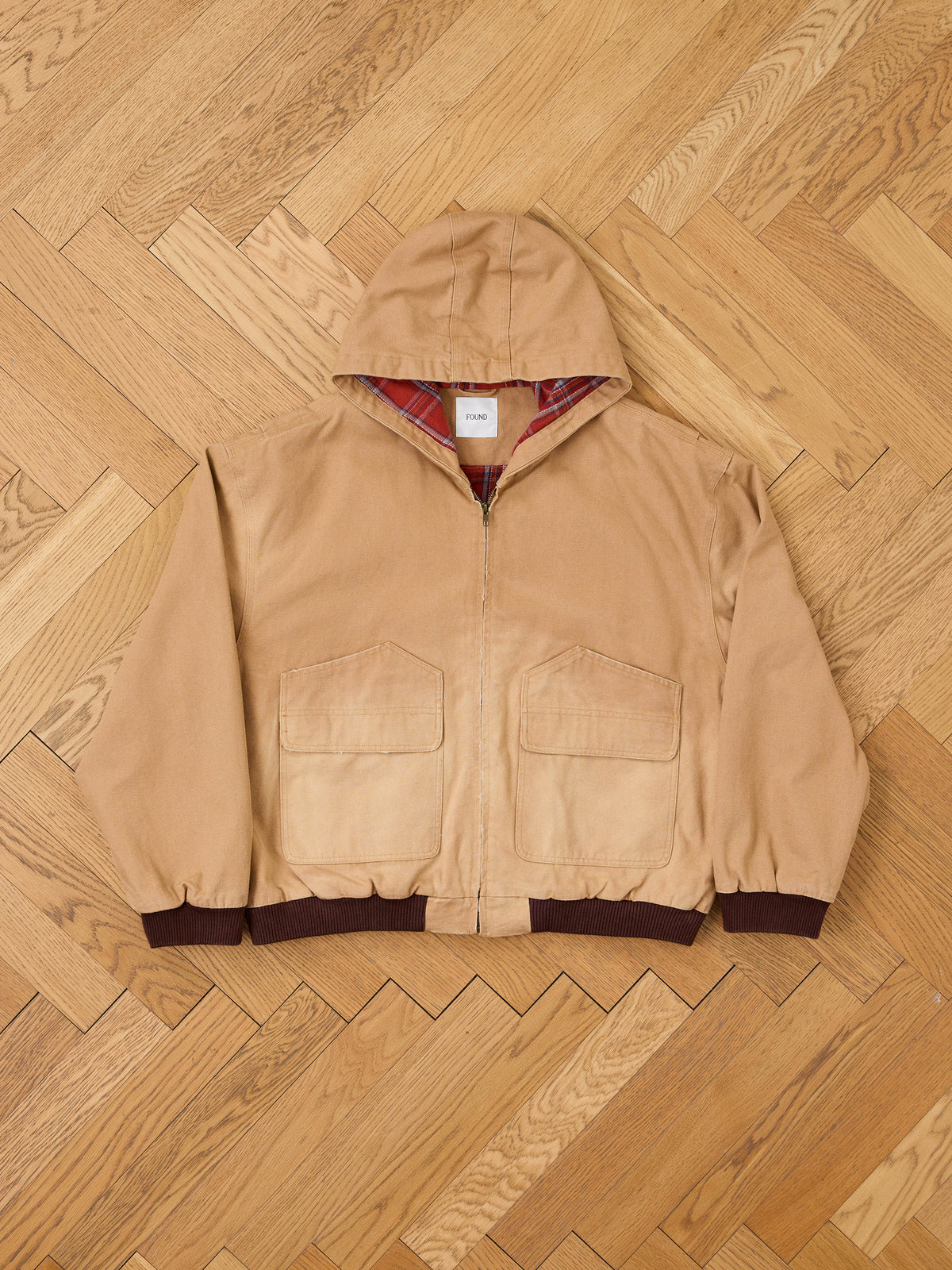 FOUND Faded Canvas Work Hoodie in tan, unisex relaxed fit, front zipper, two large pockets, dark brown cuffs, laid flat on a herringbone floor.