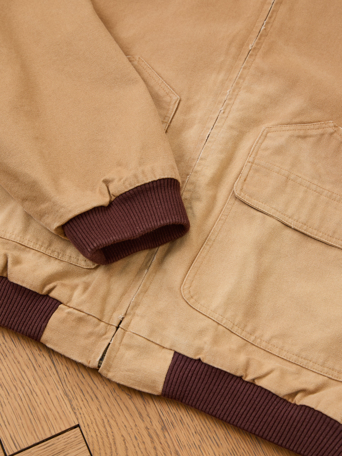 Close-up of the FOUND Faded Canvas Work Hoodie featuring a tan unisex relaxed fit, large front pockets, and brown ribbed cuffs and hem, laid flat on a wooden floor.