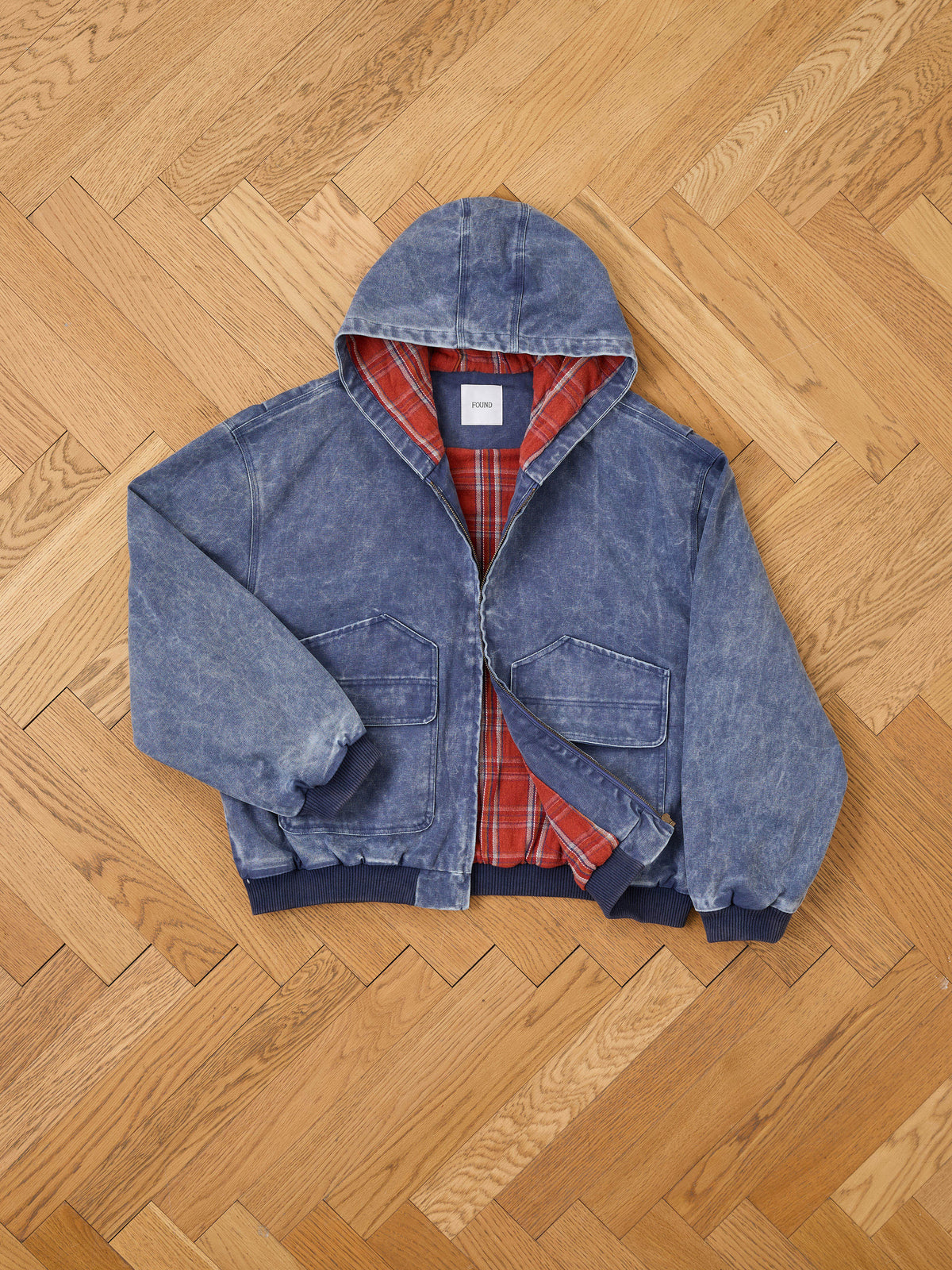 The FOUND Faded Canvas Work Hoodie, featuring a blue hooded denim design with a red plaid lining reminiscent of a plaid-lined hoodie, includes two large front pockets and ribbed cuffs and hem, displayed on a light wooden herringbone floor.