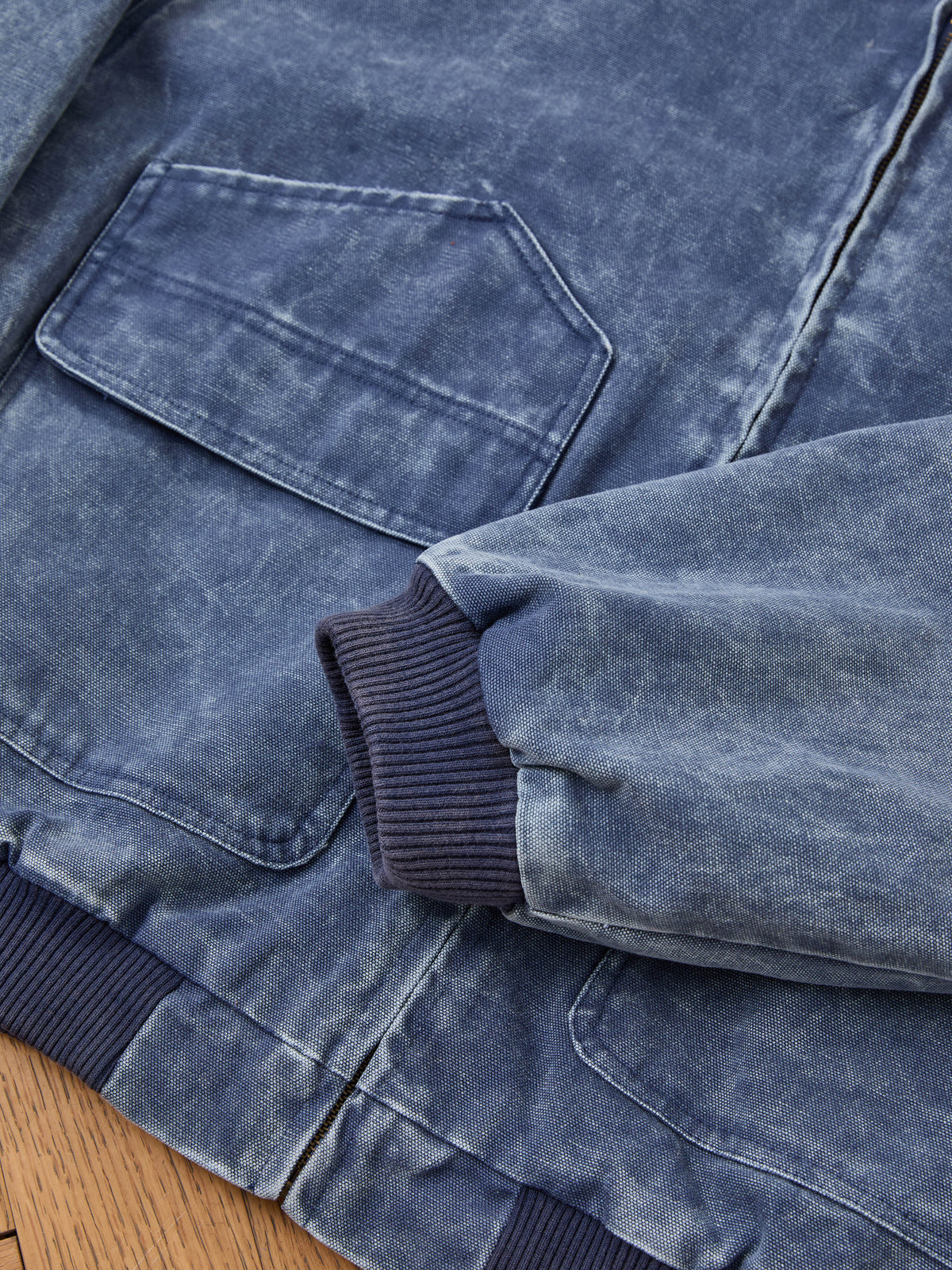 Close-up of the FOUND Faded Canvas Work Hoodie in blue, featuring a large front pocket and ribbed cuffs, laid flat on a wooden surface.
