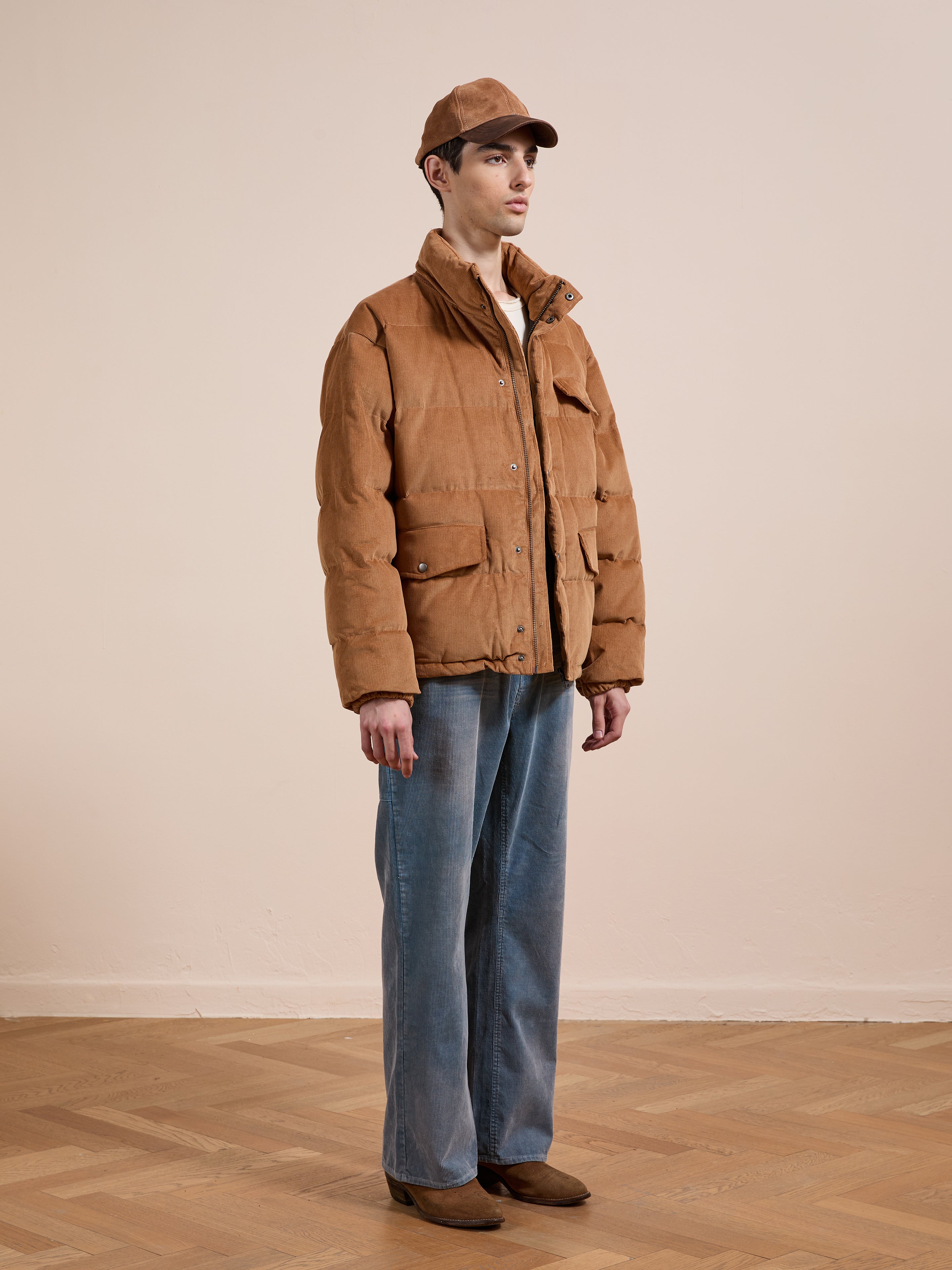 Indoors, a person wears the FOUND Corduroy Puffer Jacket in tan with a matching cap, blue wide-leg jeans, and brown shoes, standing on a wooden floor against a plain beige wall.