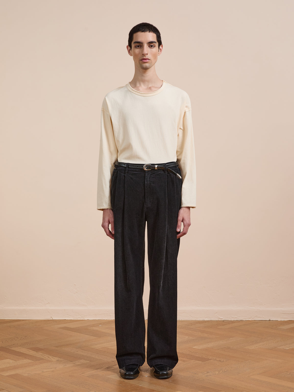 A person stands indoors on a wooden floor, facing the camera against a beige background, wearing a long-sleeve cream shirt and FOUND Faded Pleated Corduroy Pants, styled with a belt and black shoes.