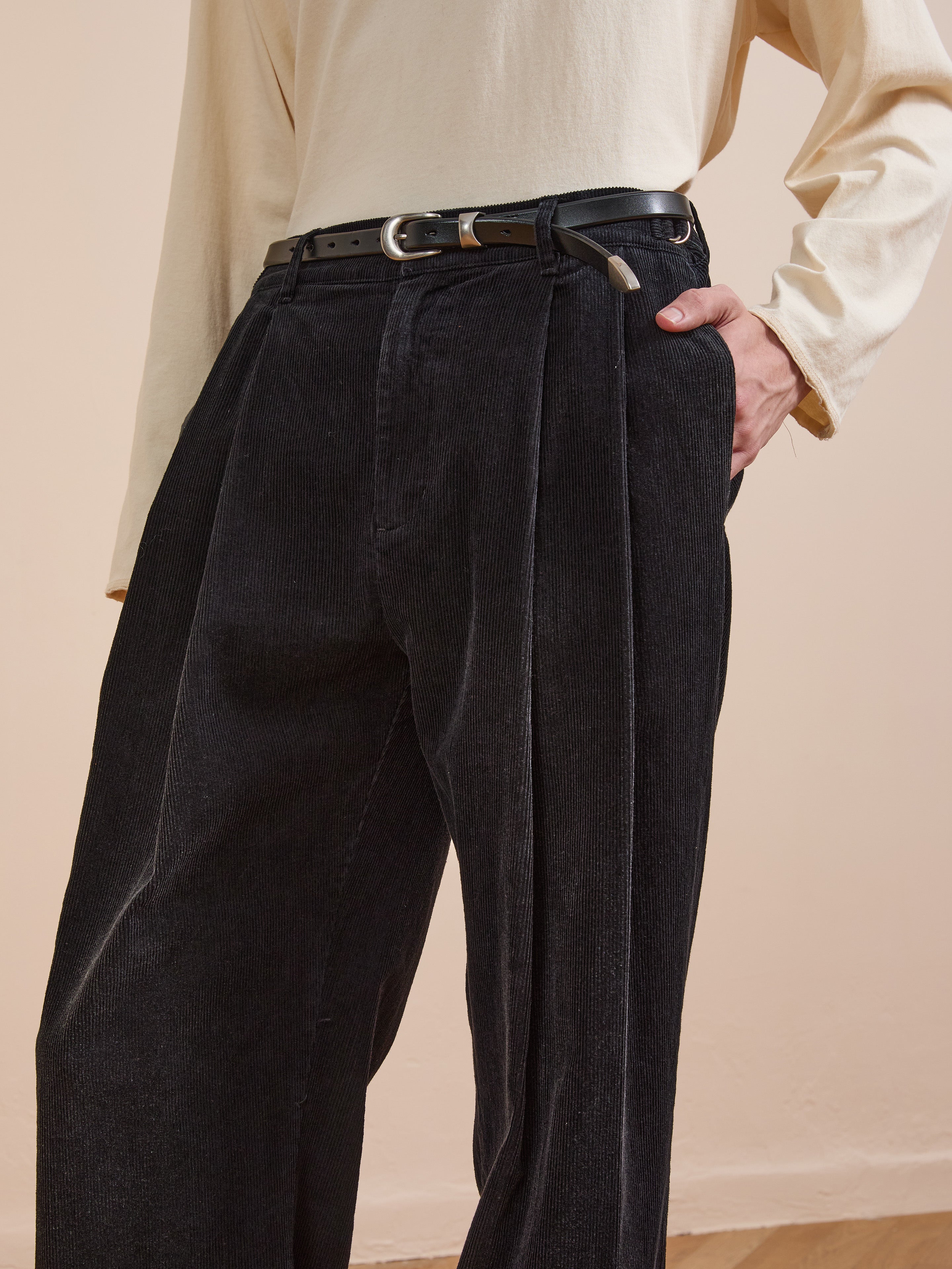 Person wearing a long-sleeved beige shirt, FOUND Faded Pleated Corduroy Pants with a wide leg relaxed fit, and a black belt with a keychain, one hand in their pocket.