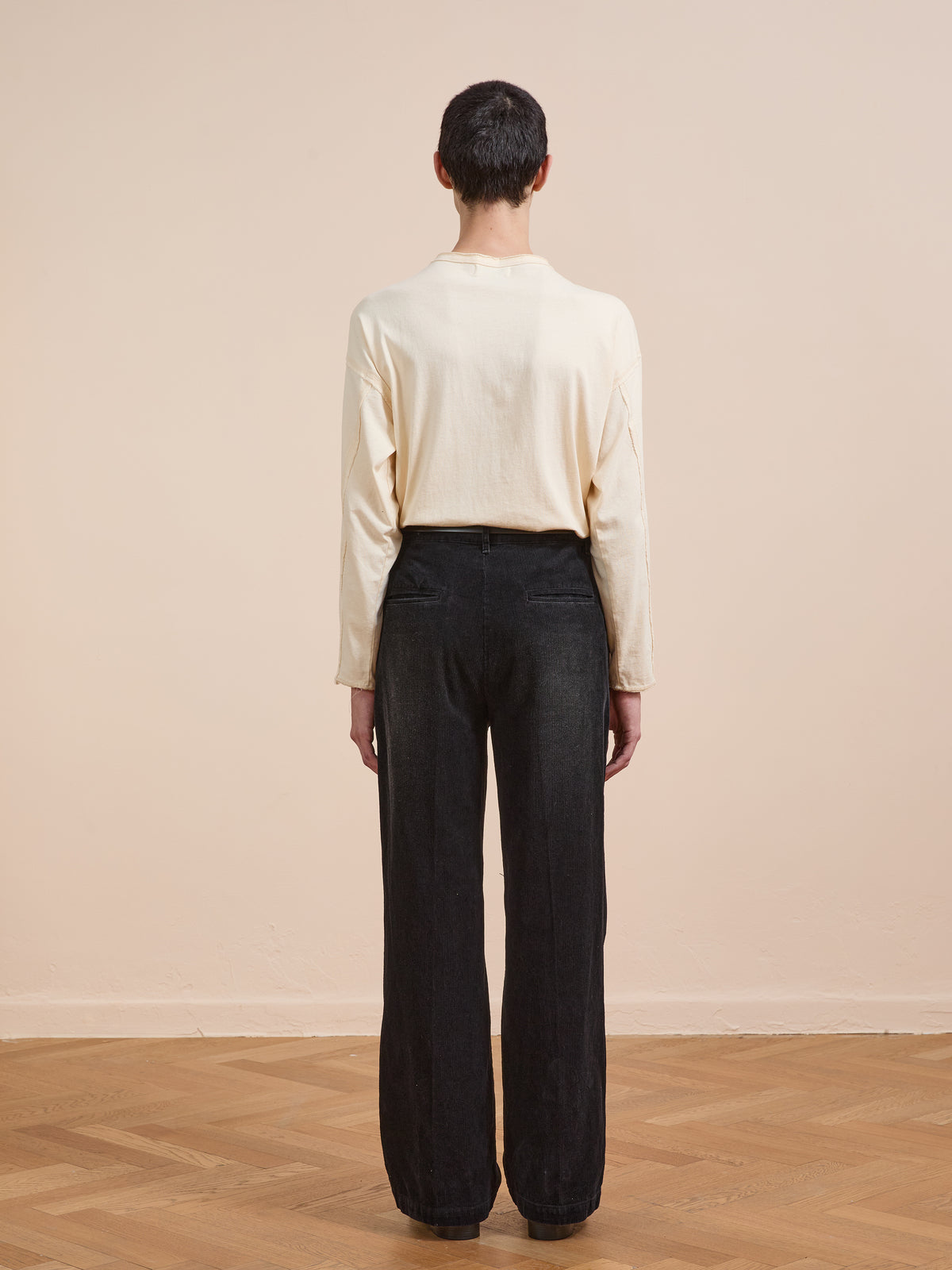 Person stands indoors with their back to the camera, wearing FOUND Faded Pleated Corduroy Pants in black, paired with a long-sleeve cream shirt on a wooden floor against a plain beige wall.