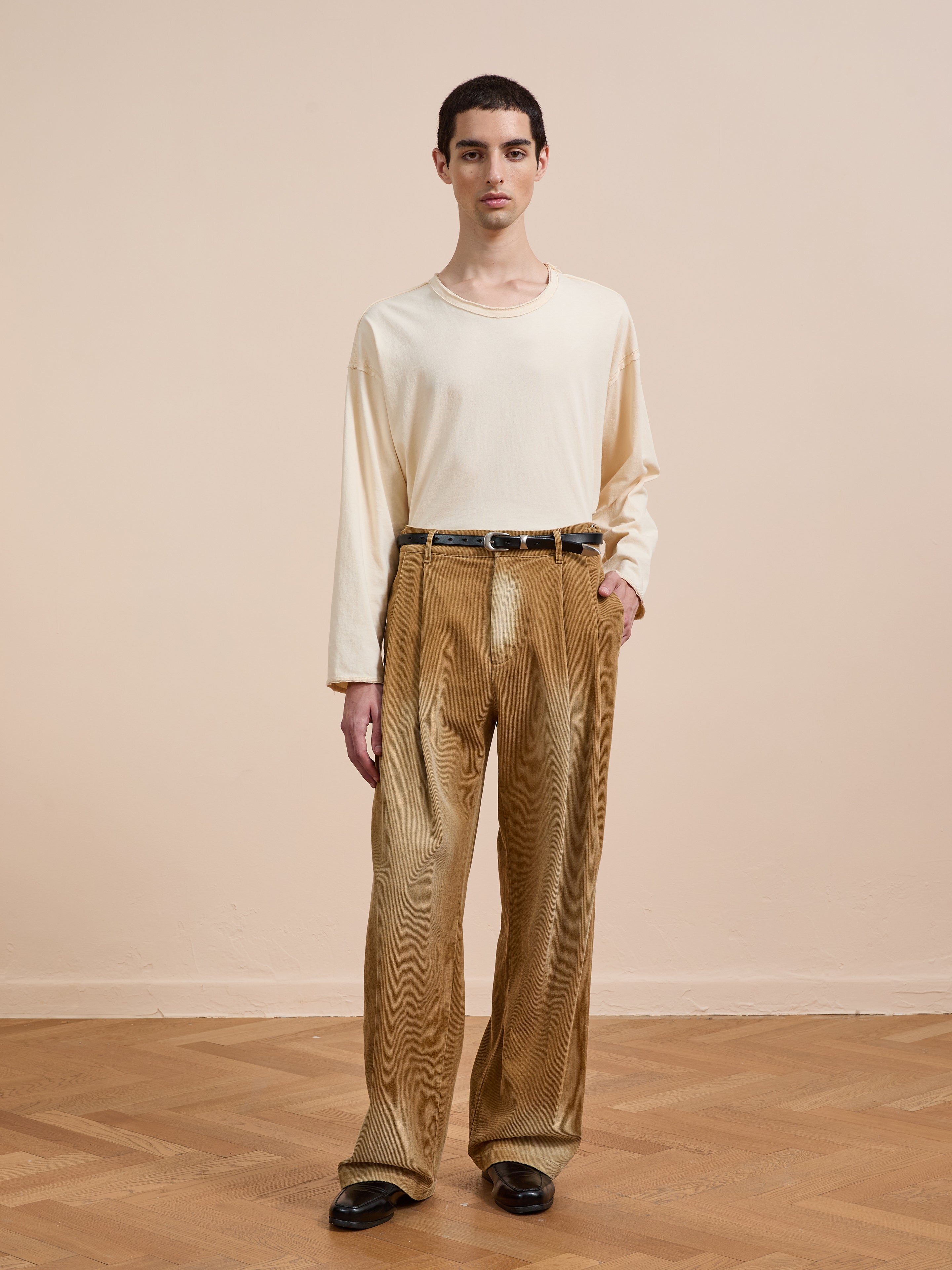 Indoors on a wooden floor, a person wears FOUND's Faded Pleated Corduroy Pants—high-waisted with double pleats and a relaxed fit—paired with a cream long-sleeve shirt, black belt, and black shoes.