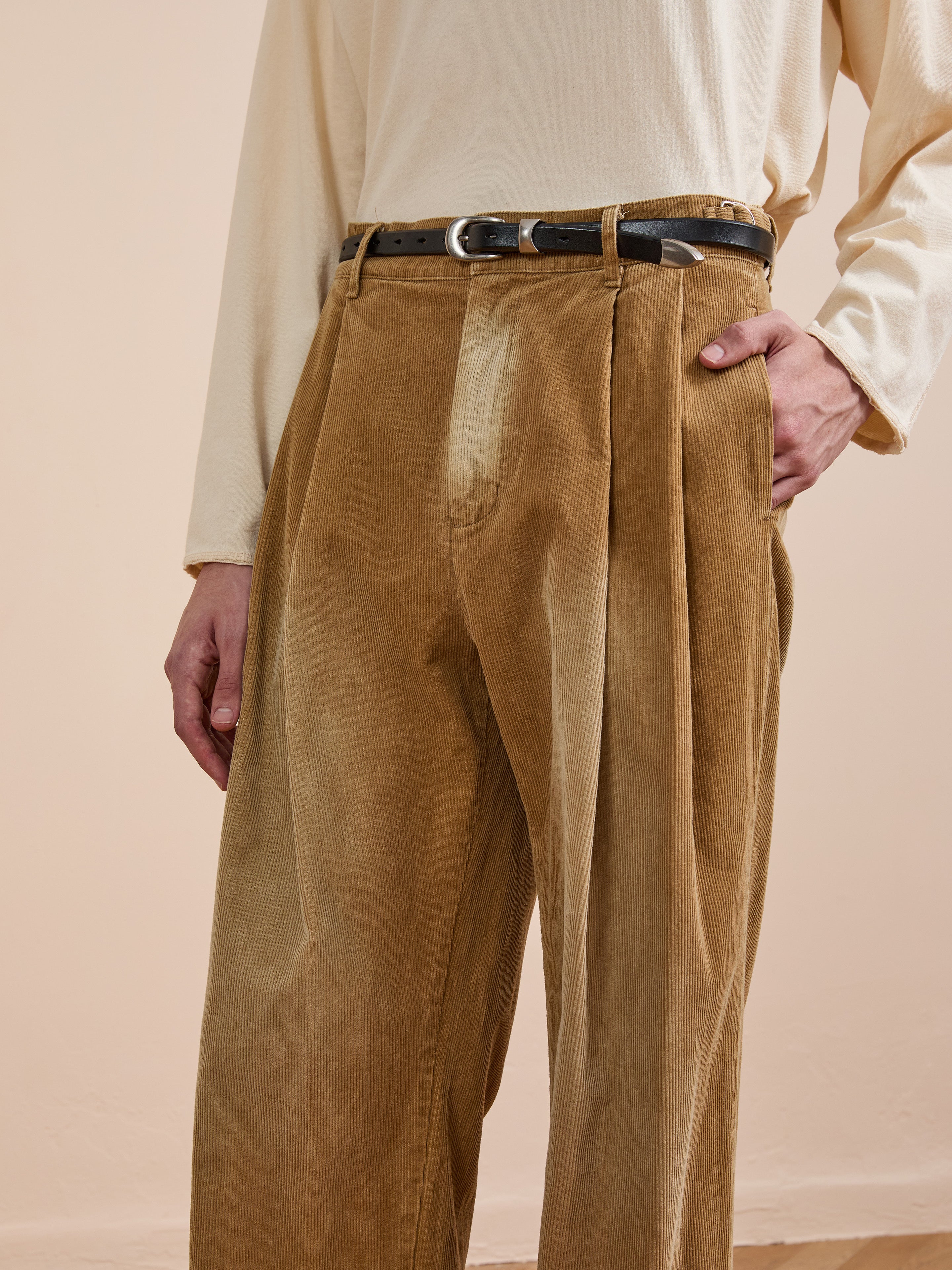Model wears FOUND Faded Pleated Corduroy Pants in tan with a cream long-sleeve shirt and black belt, hand in pocket, shown from shoulders to mid-thigh against a neutral background.