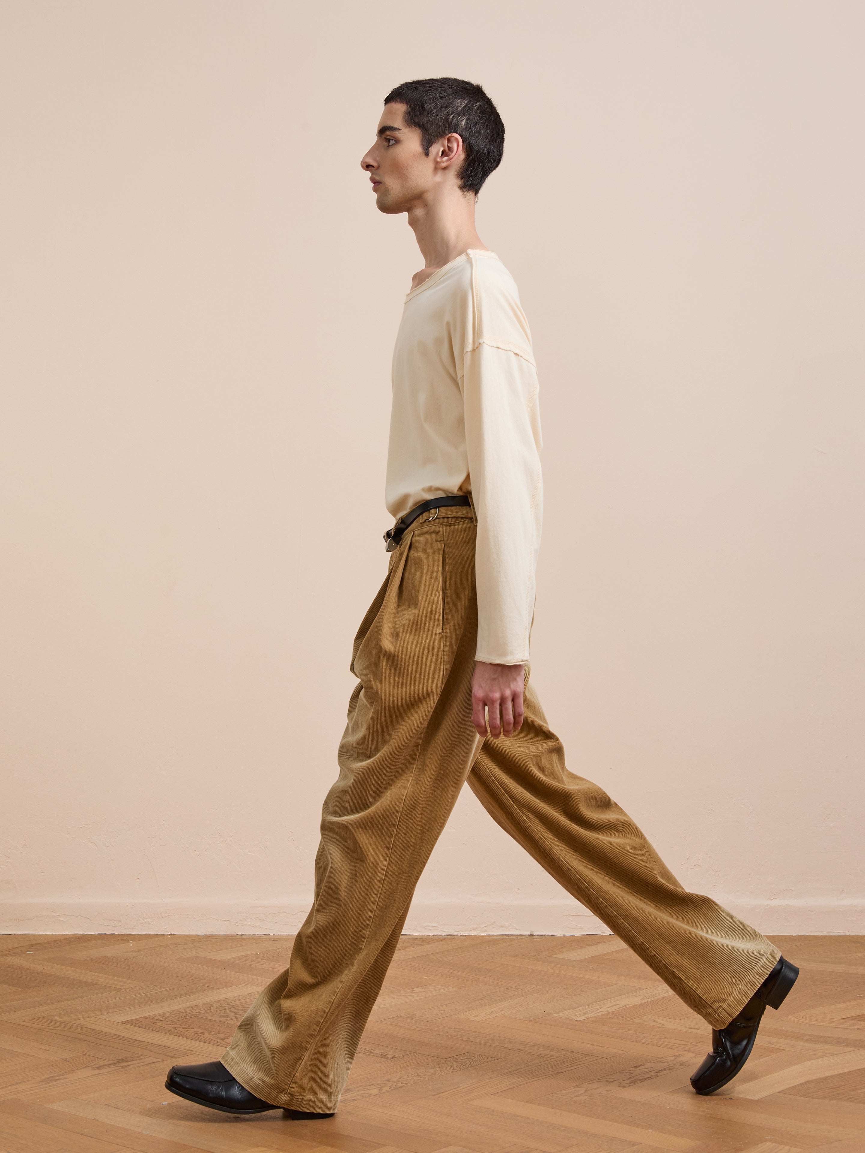 A person with short dark hair, in a cream long-sleeve shirt and FOUND's Faded Pleated Corduroy Pants, walks indoors on a wooden floor against a beige wall.
