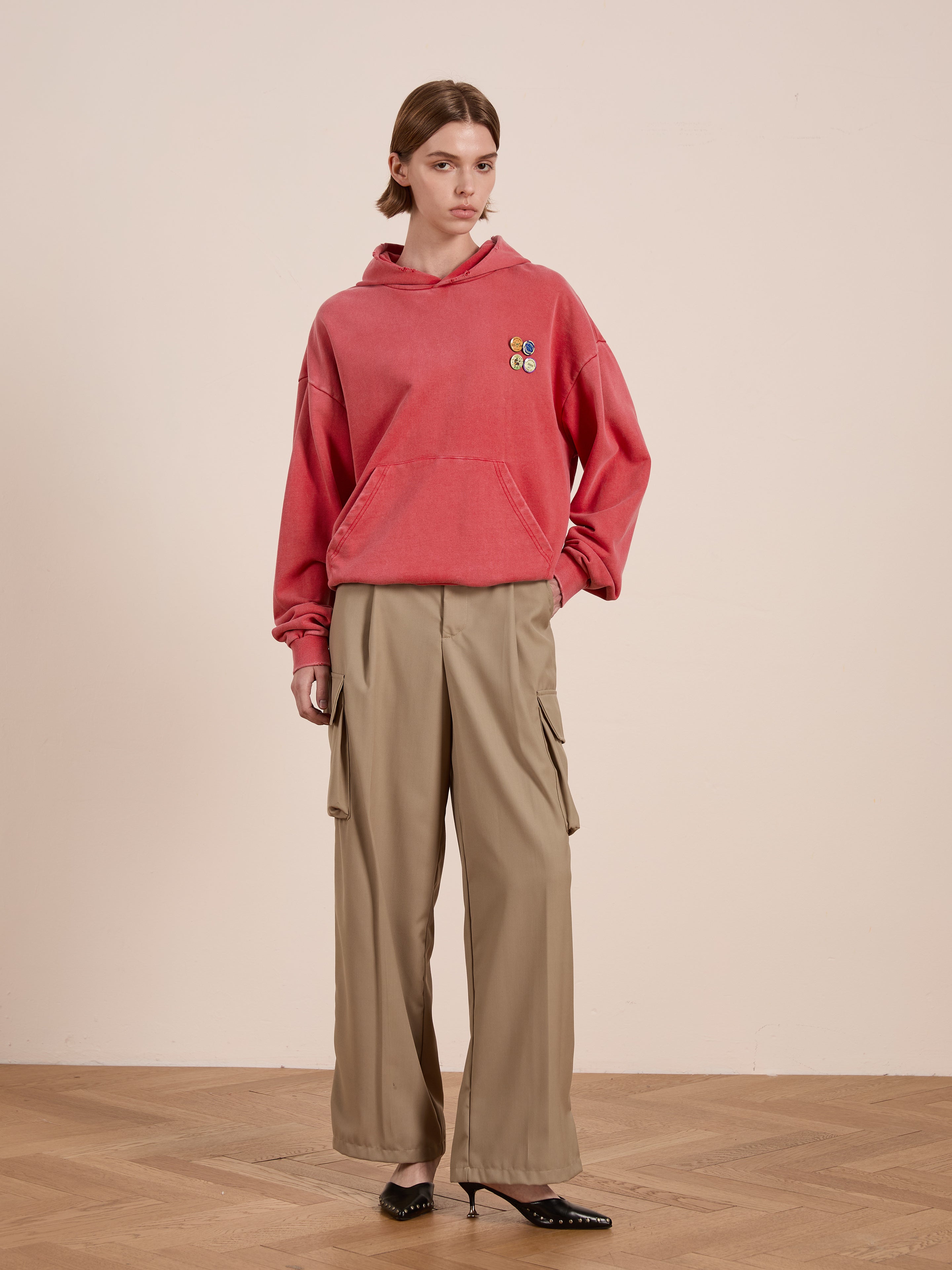 A person in a vintage-inspired oversized Multi Patch Hoodie by FOUND, beige cargo pants, and black shoes stands on a wooden floor against a plain wall.