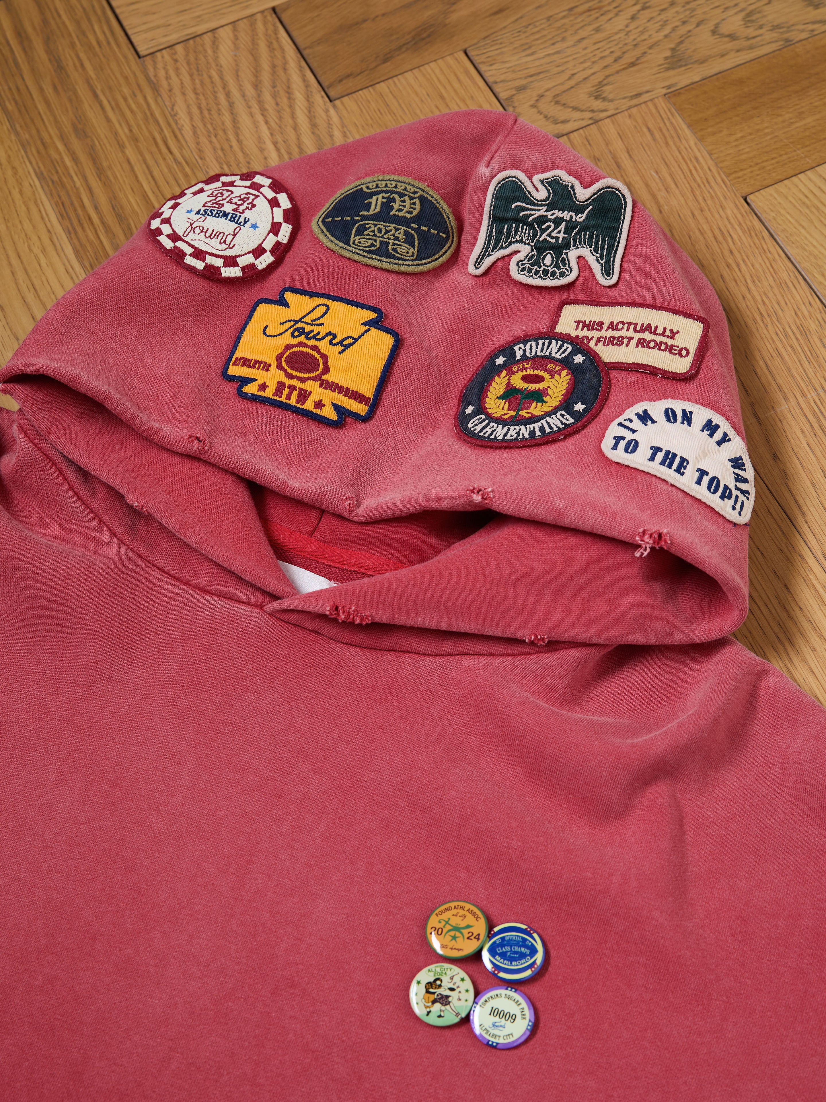 The FOUND Multi Patch Hoodie, featuring a sun-faded red wash and vintage look with colorful patches and pins on the hood including eagles, messages, and emblems, is displayed on a wooden floor.