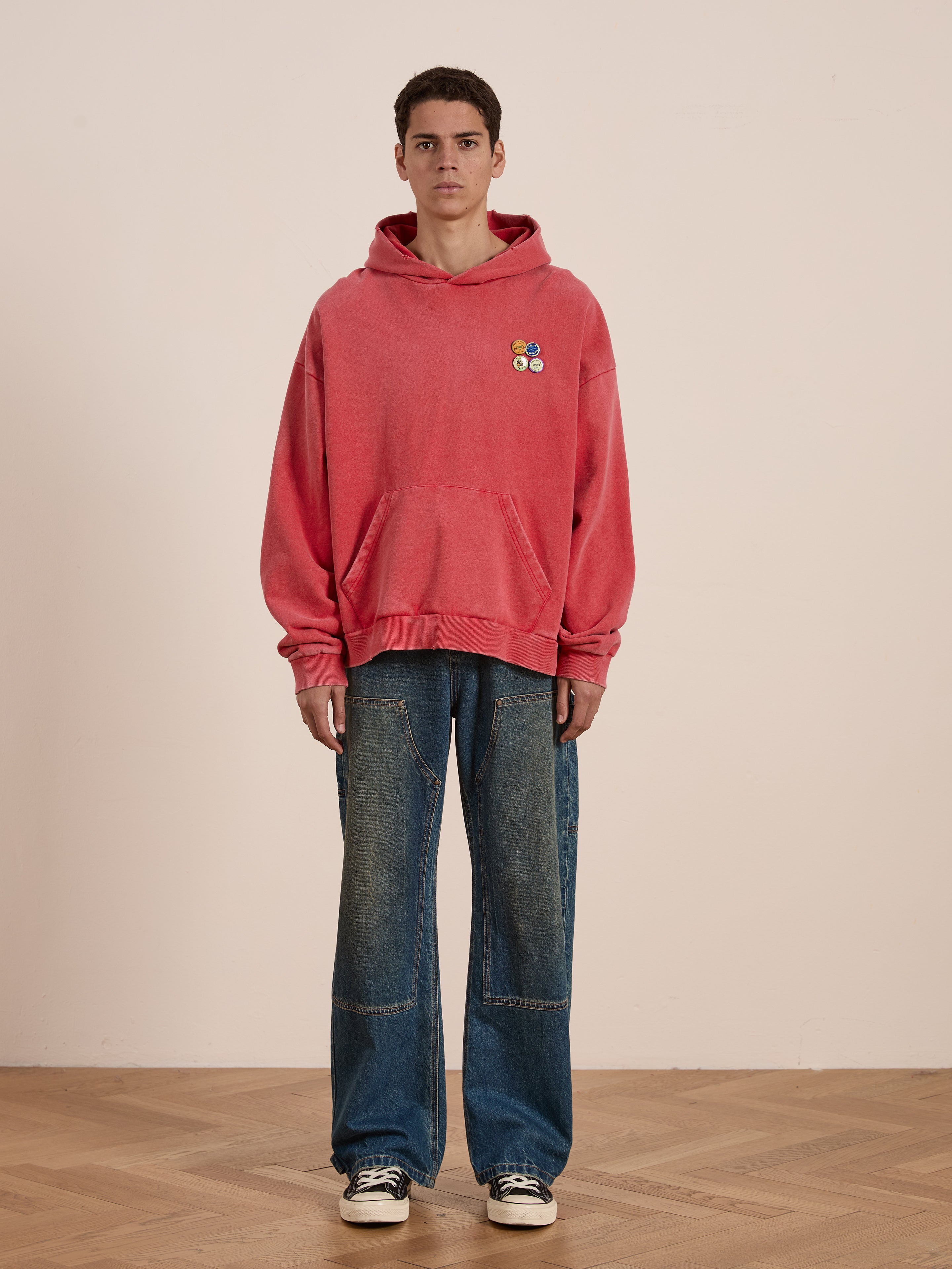 A person confidently stands on a wooden floor, wearing FOUND's Multi Patch Hoodie and blue wide-leg jeans with a sun-faded wash.