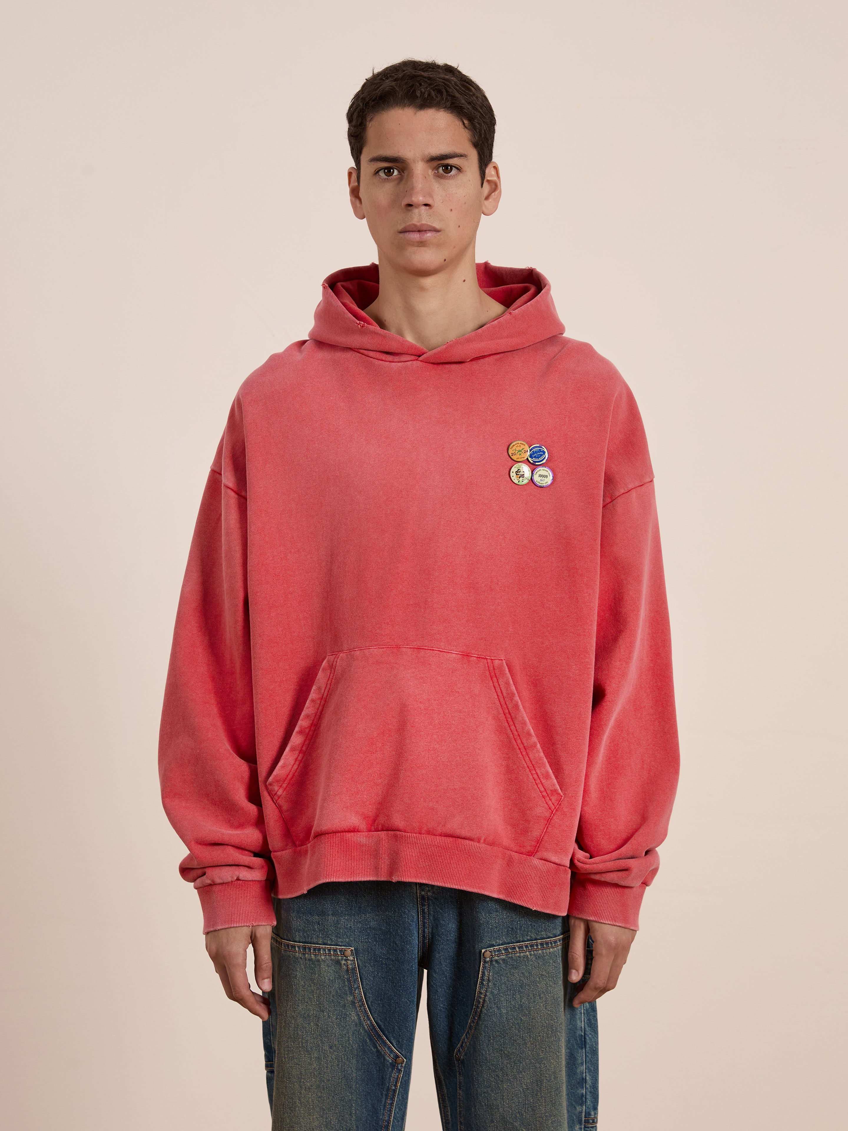 Against a plain background, a person wears FOUND's Multi Patch Hoodie in red, featuring vintage-inspired appliqués and smiley patches. Paired with blue jeans, the look is casual yet playful.