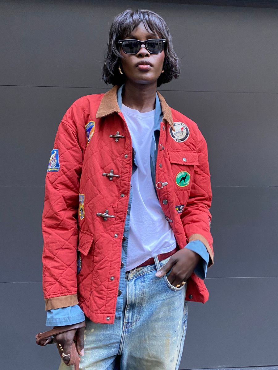 A person with sunglasses is wearing the Farmstead Quilt Patch Jacket by Found in red, over a blue denim shirt and white tee, paired with faded jeans.