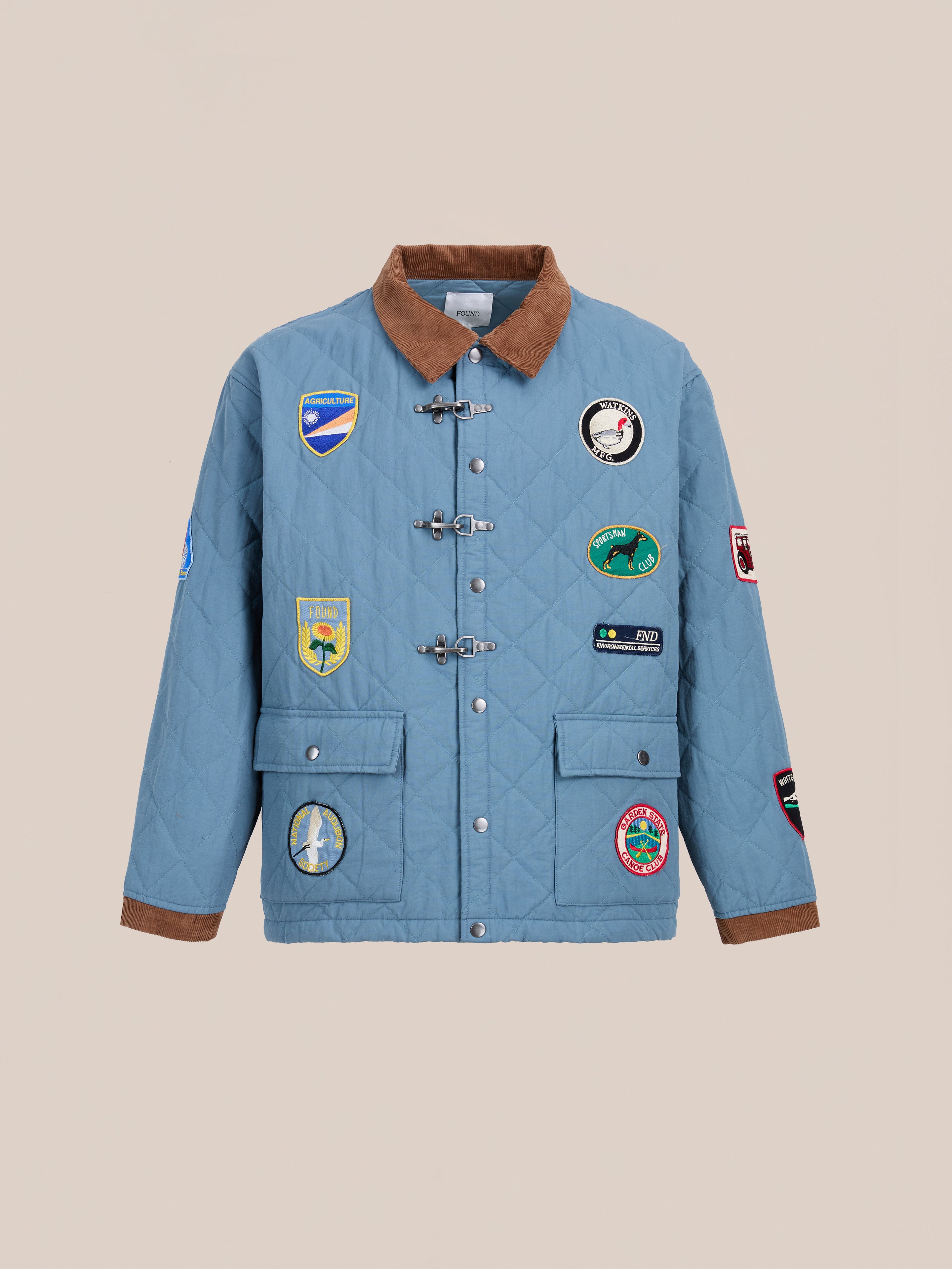 The FOUND Farmstead Quilt Patch Jacket features a light blue quilted design with a brown collar and cuffs, toggle buttons, front pockets, and colorful embroidered patches on the front and sleeves for a vintage-inspired look.