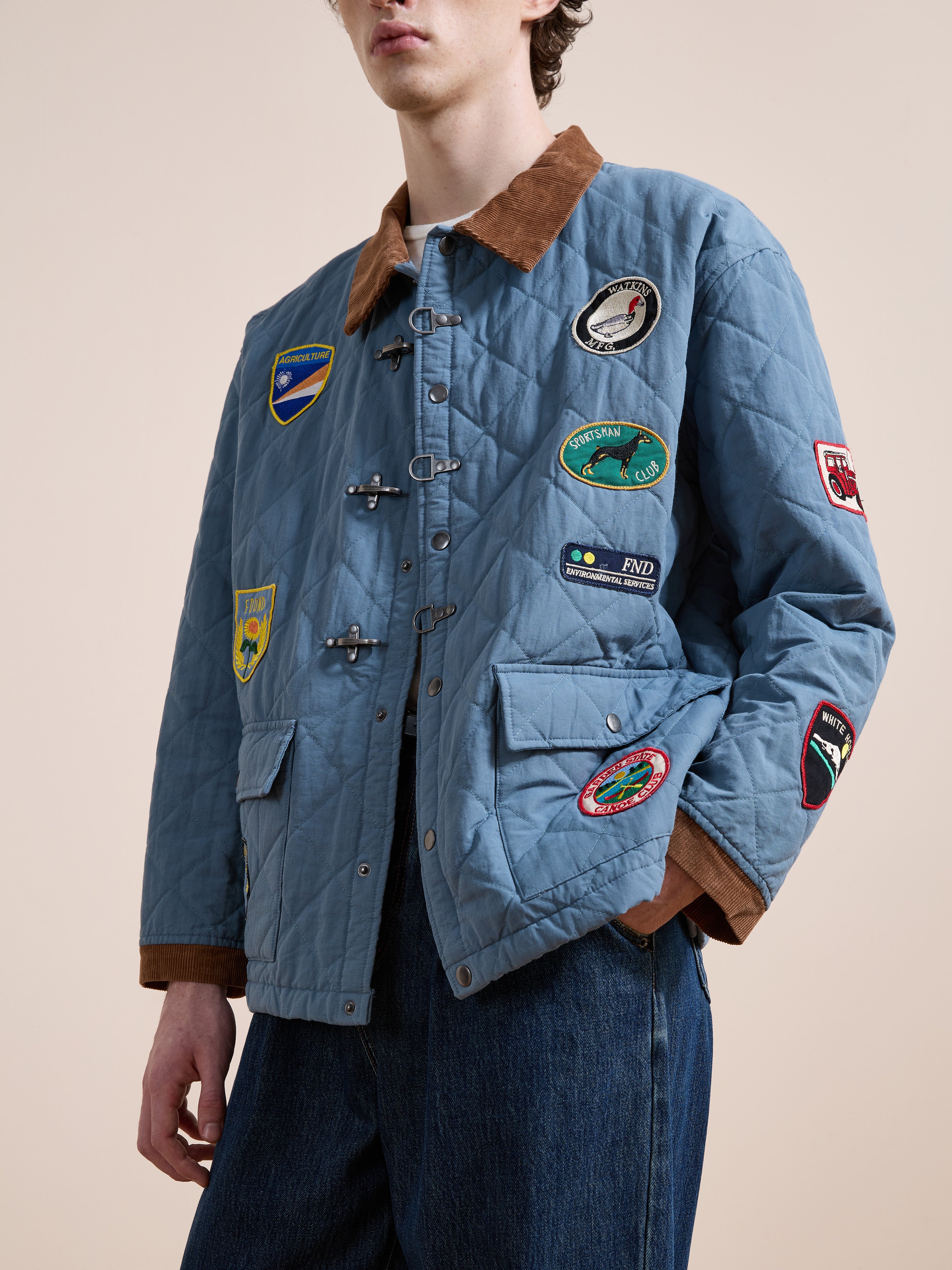 A person wears the FOUND Farmstead Quilt Patch Jacket—a blue quilted style with a brown collar and colorful patches—paired with blue jeans, standing against a plain background.