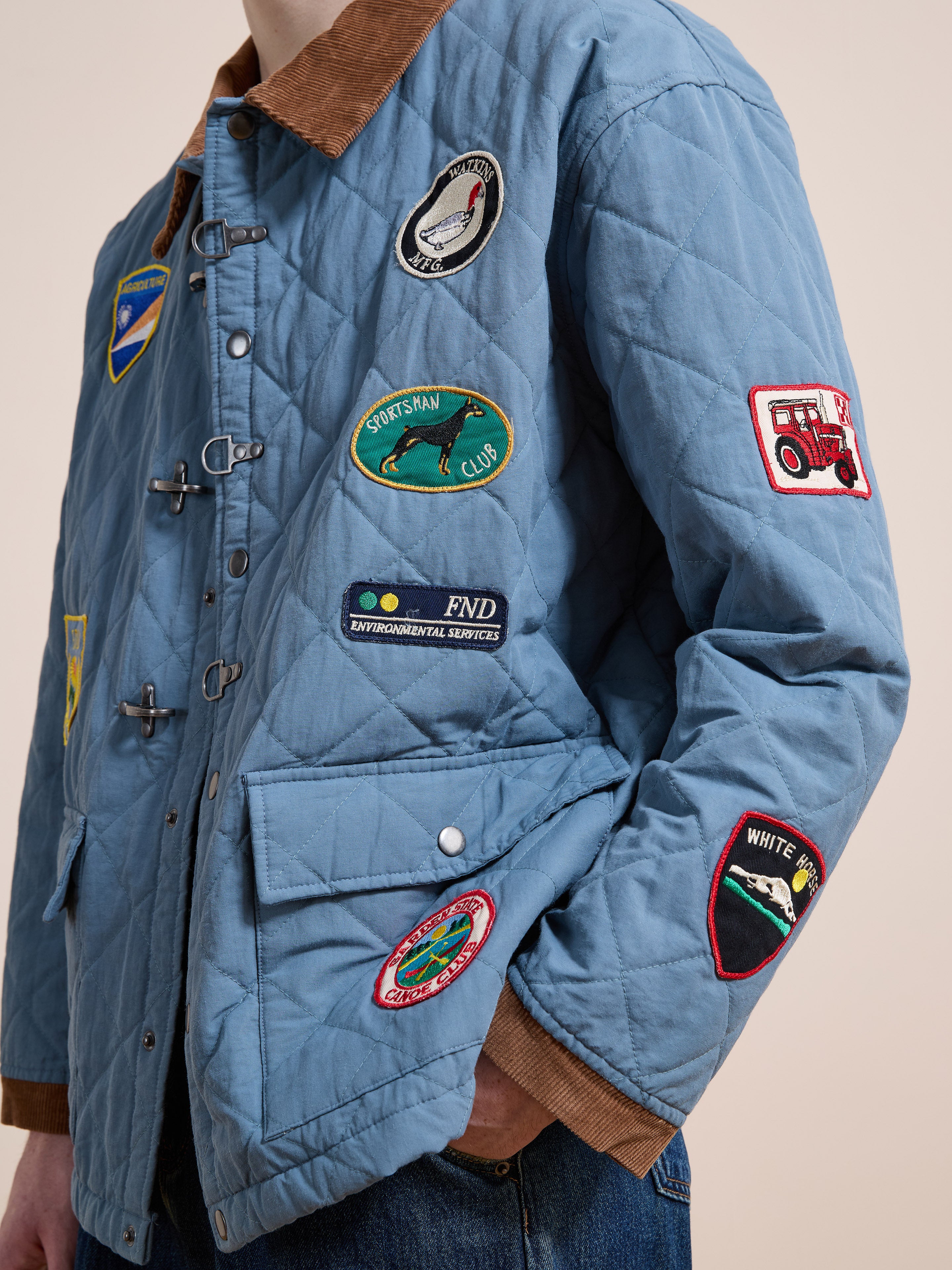 Someone wears the FOUND Farmstead Quilt Patch Jacket in light blue, featuring a brown collar and multiple colorful embroidered patches on the front and sleeve.